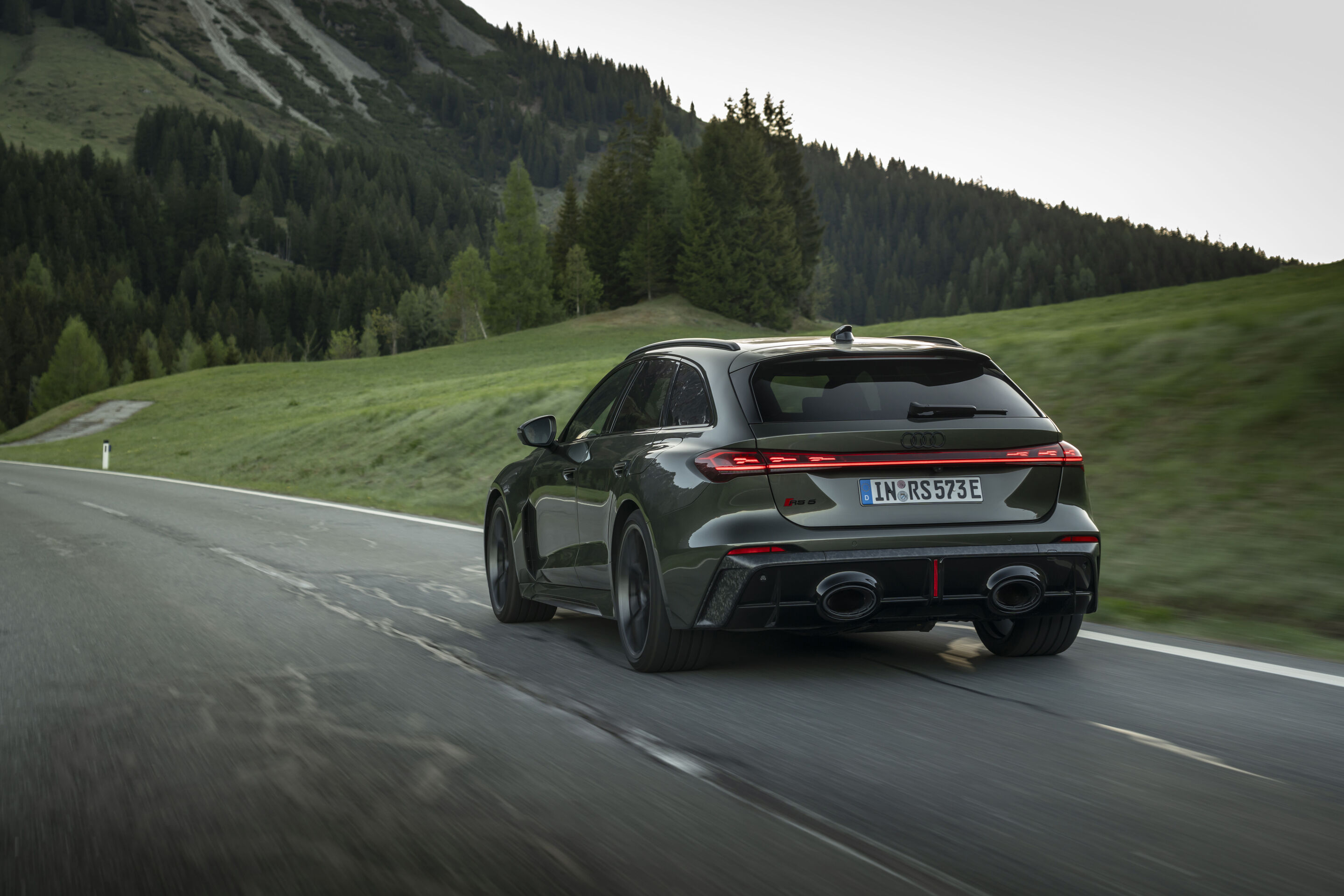 Audi RS 5 Avant - Audi RS 5 Avant in Bedfordgrün, Outdoor-Fahraufnahme, Exterieur, Heckansicht auf kurvenreicher Landstraße in bergiger Landschaft.