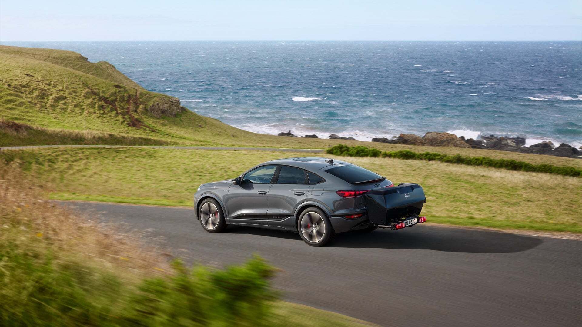 Ein Audi Q6 Sportback e-tron auf einer Straße direkt am Meer. Die Fläche neben der Straße sind grün bewachsen.