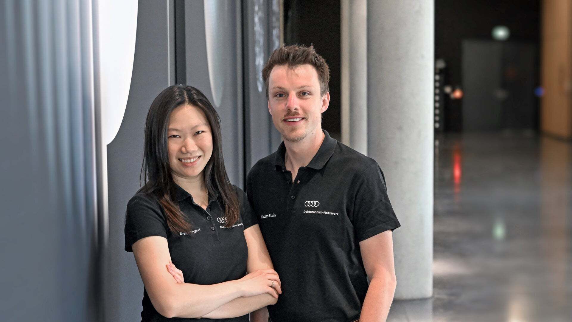 Two smiling people wearing black Audi polo shirts are standing in a modern interior.