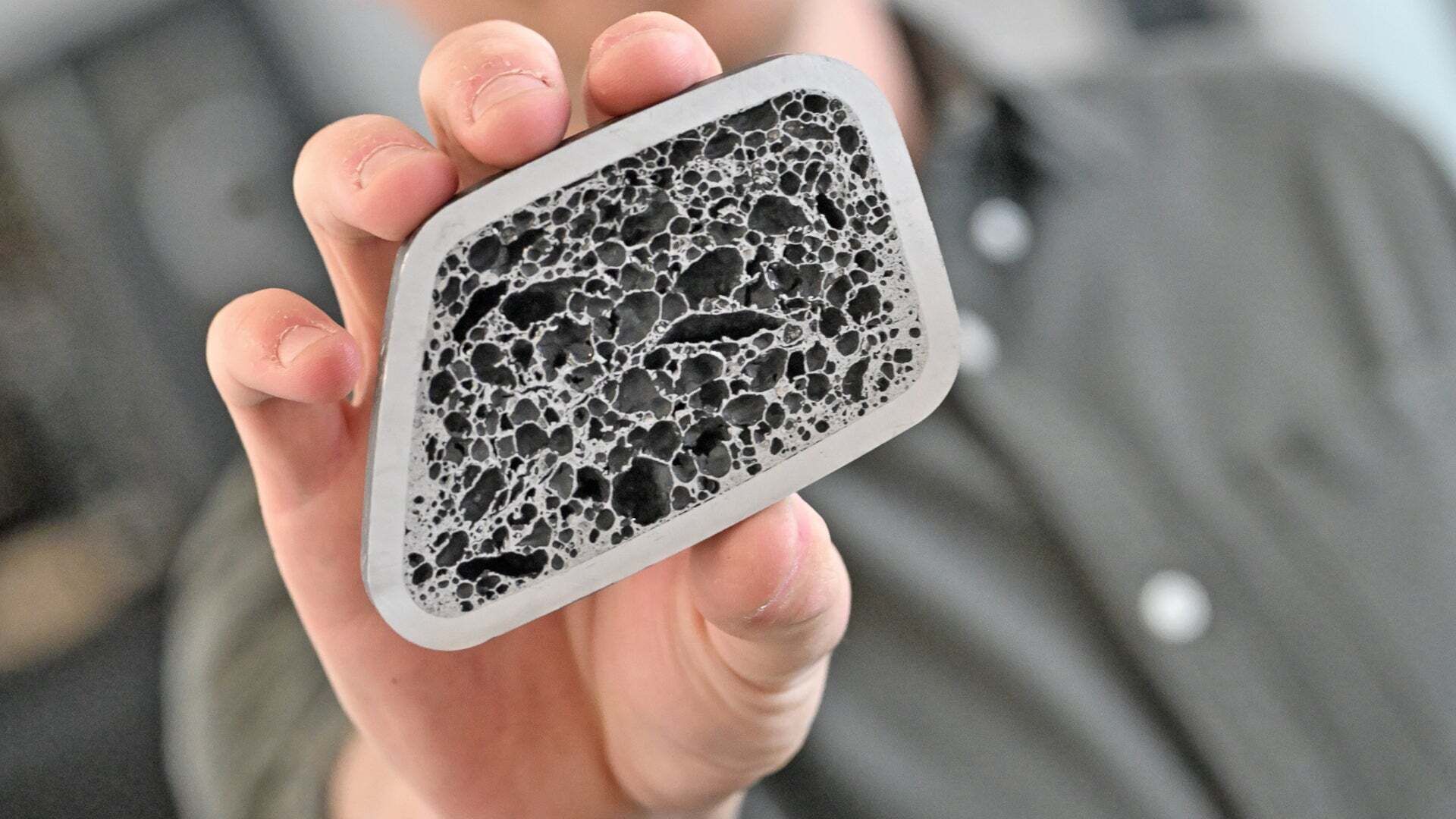 Hand hält eine rechteckige Materialprobe mit poröser schwarz-grauer Struktur.