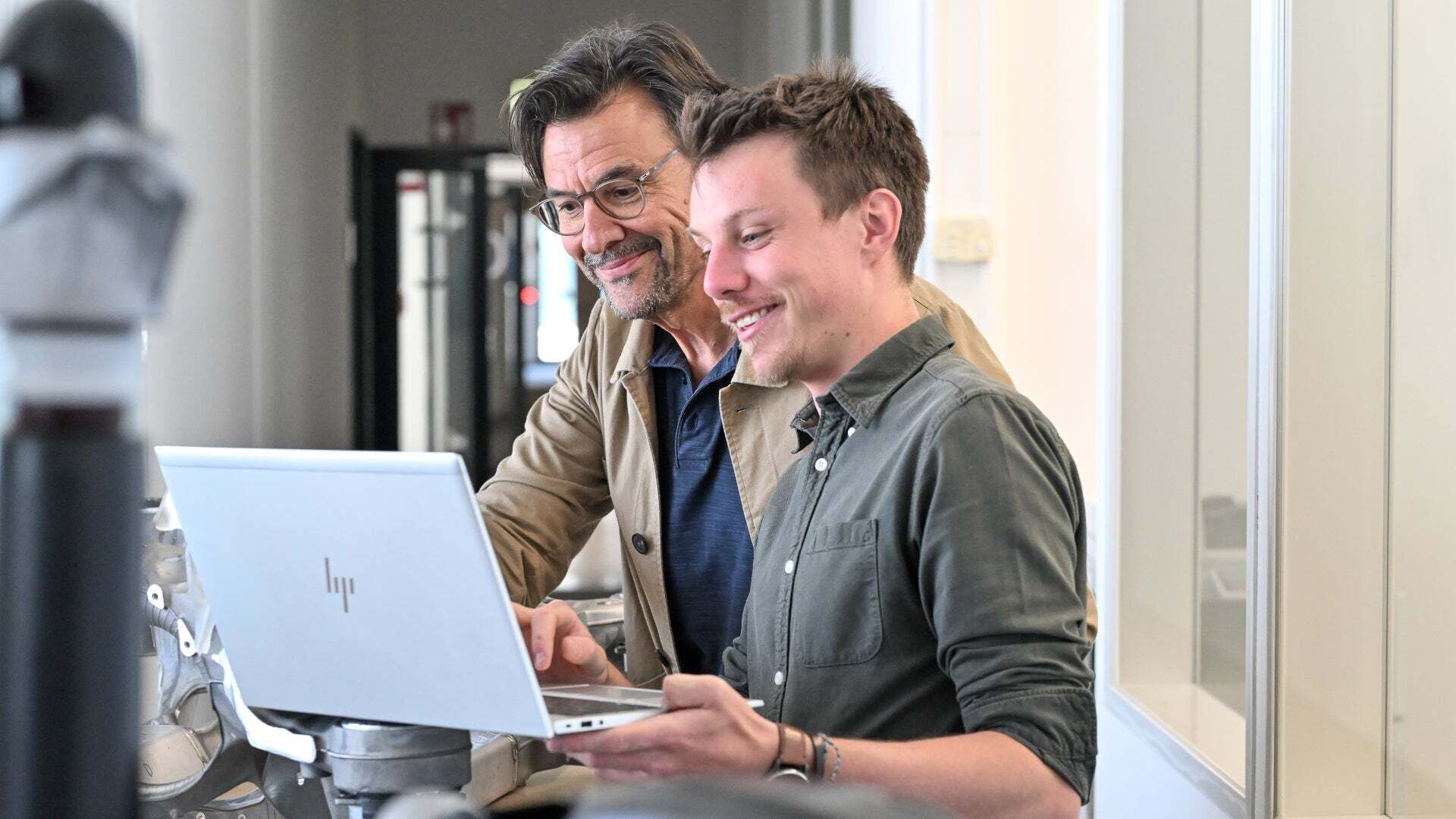 Zwei Männer schauen gemeinsam auf einen Laptop in einem technischen Arbeitsbereich.