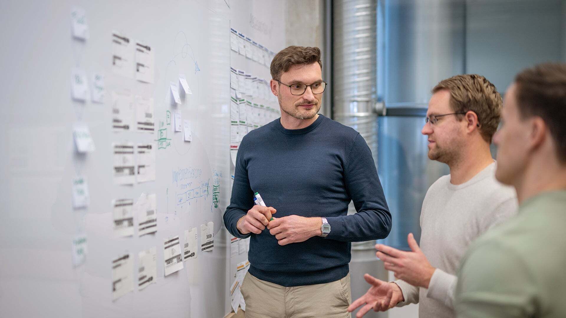 Alexander zeigt zwei Kollegen etwas an einem Whiteboard