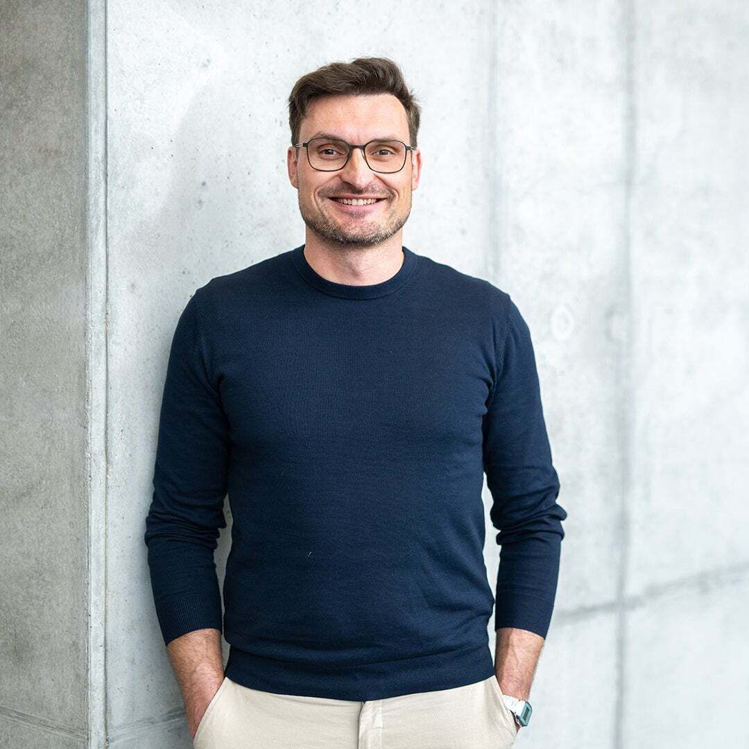 Alexander Lang hat die Hände in den Hosentaschen, lächelt in die Kamera und ist an eine Wand gelehnt