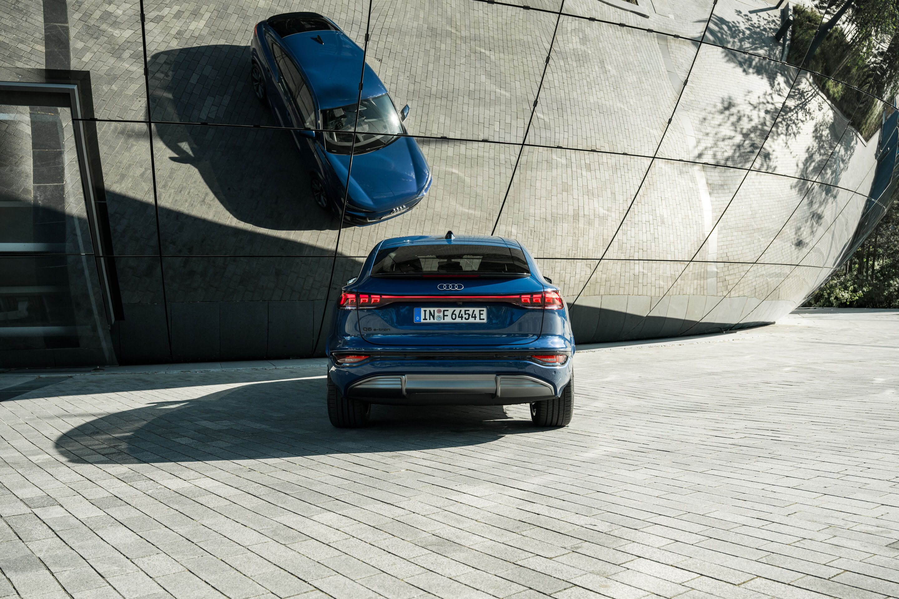 Audi Q6 e-tron Lifecycle Experience - Standaufnahme: Audi Q6 e-tron (Ascariblau), Heckansicht vor einer gebogenen, spiegelnden Architektur.
