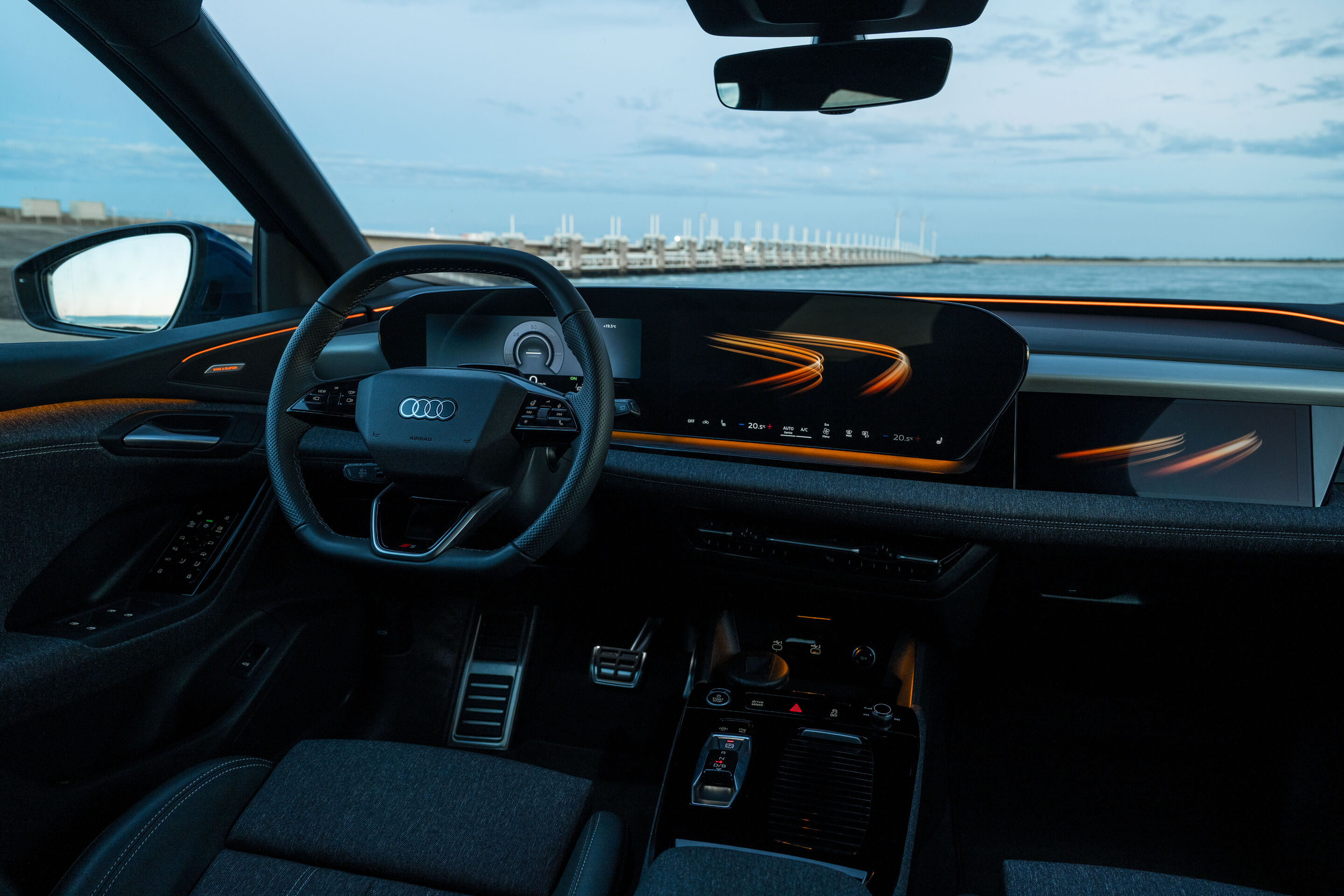 Audi Q6 e-tron Lifecycle Experience - Innenraum des Audi Q6 e-tron mit Darstellung der Erlebniswelt: Blick auf Lenkrad, digitales Cockpit und durchgehendes Display, akzentuiert durch orange Ambientebeleuchtung