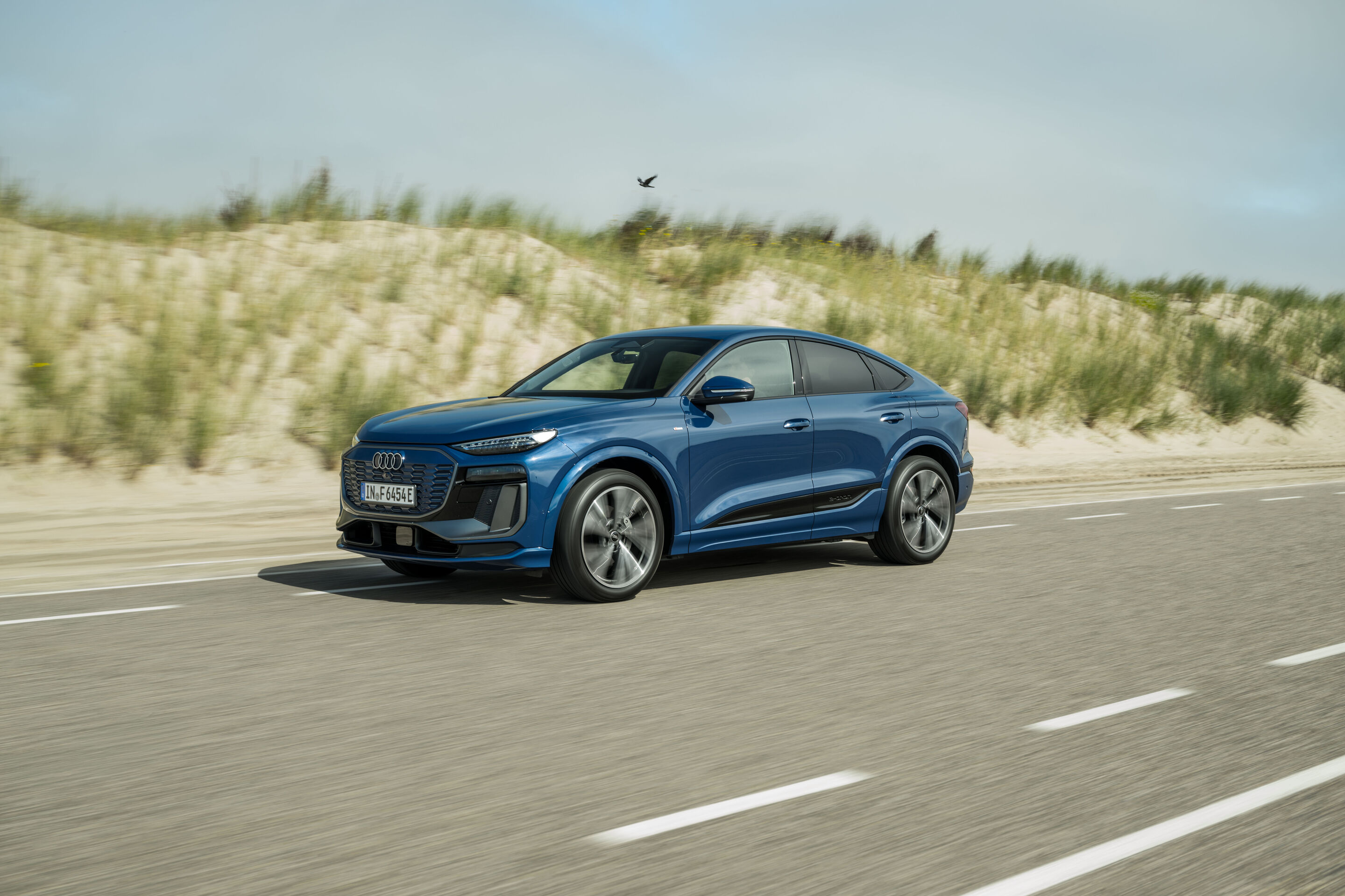 Audi Q6 e-tron Lifecycle Experience - Dynamische Aufnahme: Audi Q6 e-tron Sportback (Ascariblau), Front-Dreiviertelansicht in Bewegung auf einer Straße entlang von Dünenlandschaft