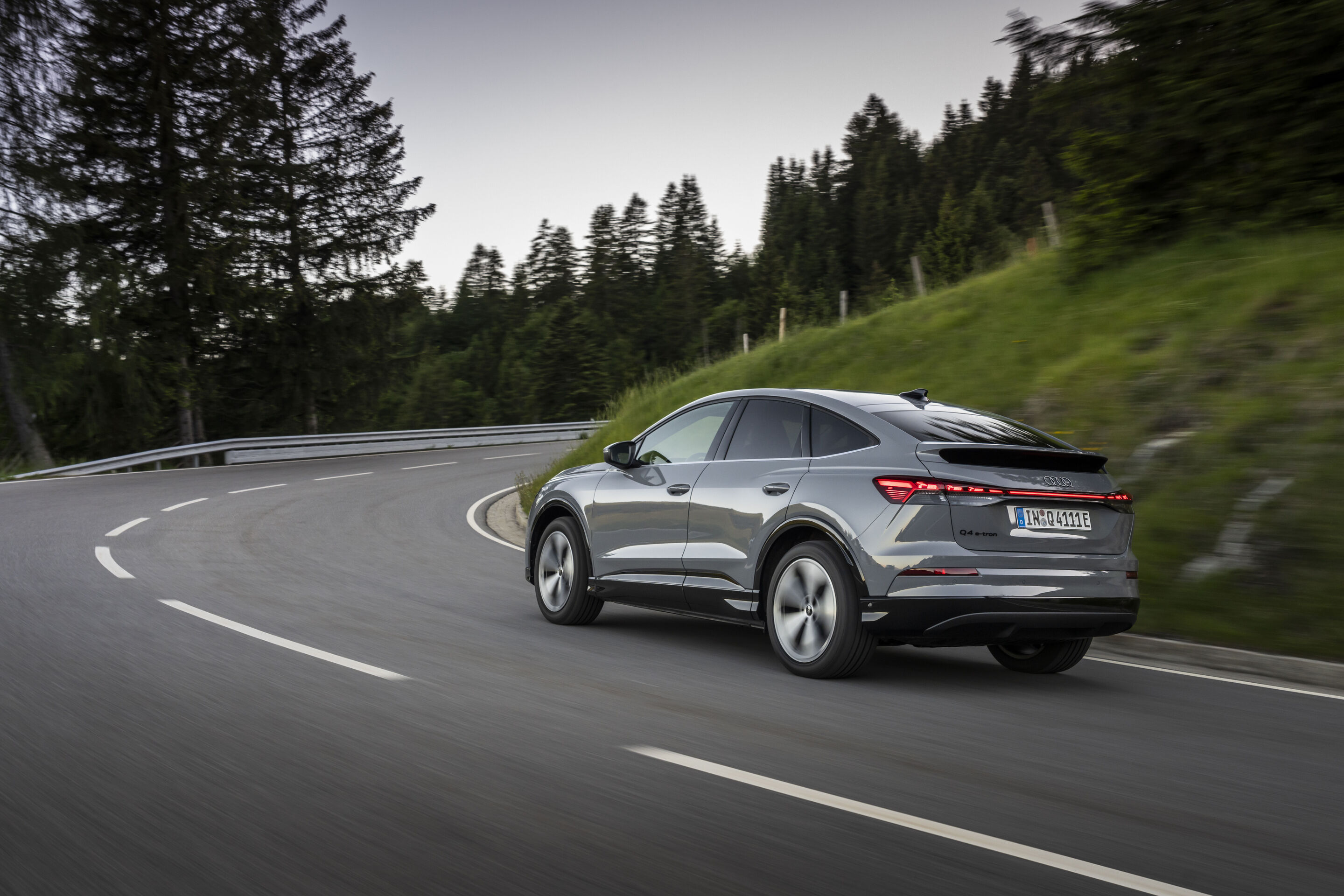Ein Auto für jeden Moment: Unterwegs im Audi Q4 e-tron - Audi Q4 Sportback e-tron in Pebblegrau, Outdoor-Fahraufnahme, Exterieur, Dreiviertelheckansicht auf kurvenreicher Straße durch bewaldete Umgebung.