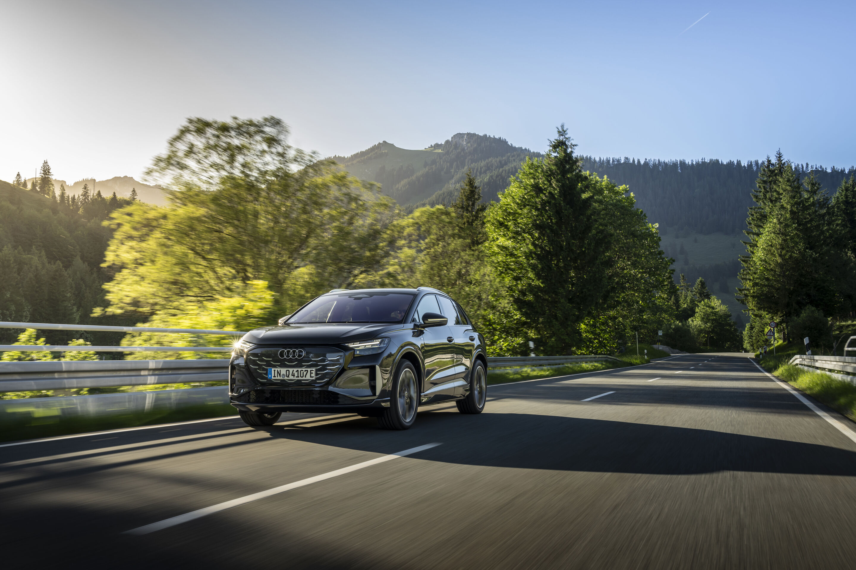 Ein Auto für jeden Moment: Unterwegs im Audi Q4 e-tron - Audi Q4 SUV e-tron in Tamboragrau, Outdoor-Fahraufnahme, Exterieur, Dreiviertelfrontansicht auf einer Landstraße, umgeben von Wald und Bergkulisse.
