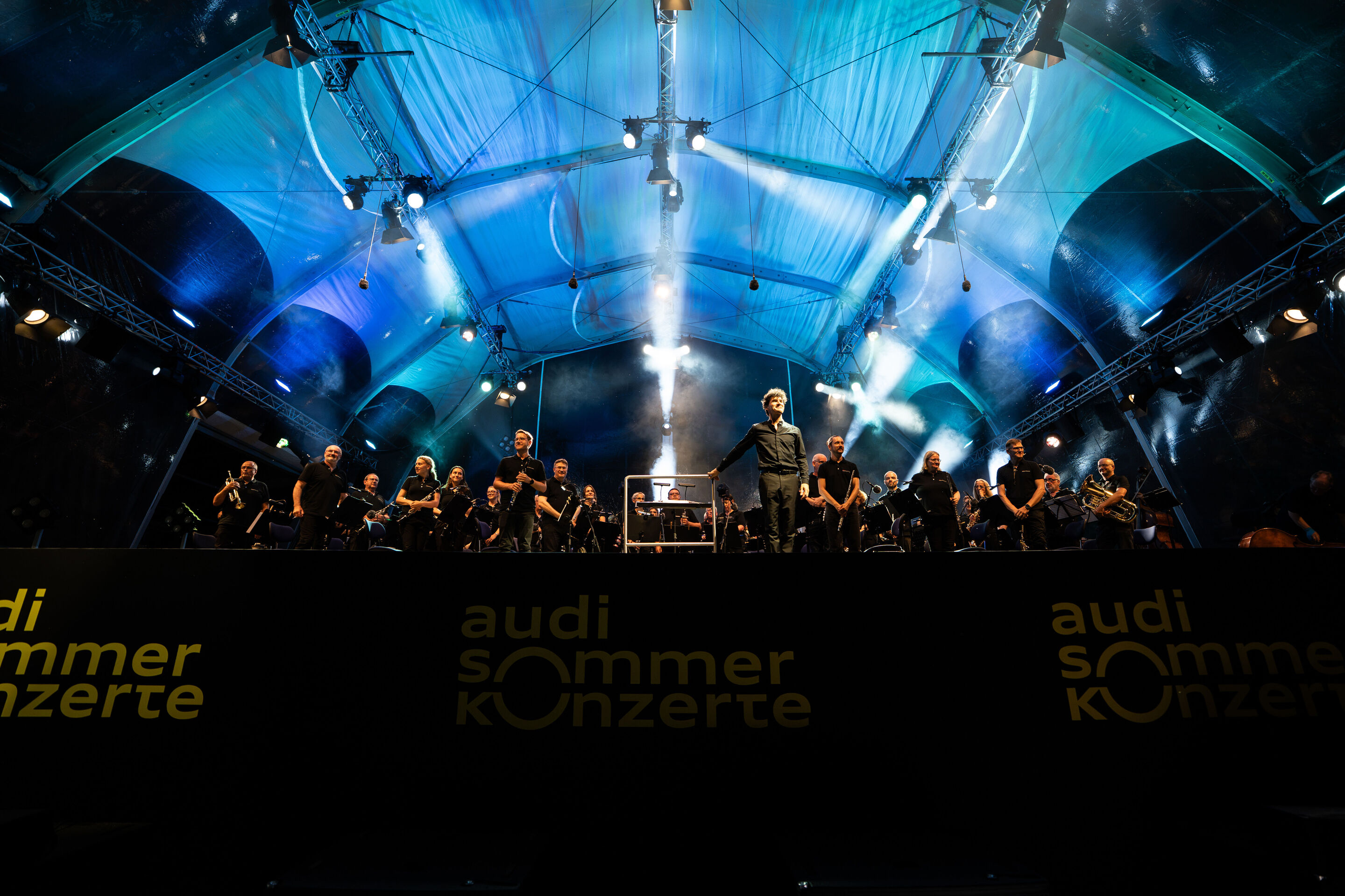 Die Audi Bläserphilharmonie beim Eröffnungskonzert/Klassik Open Air im Klenzepark - Die Audi Bläserphilharmonie auf der Bühne des Klassik Open Airs im Klenzepark 2026 mit Dirigent Mathias Wehr im Zentrum der Aufnahme vor den Musikerinnen und Musikern...