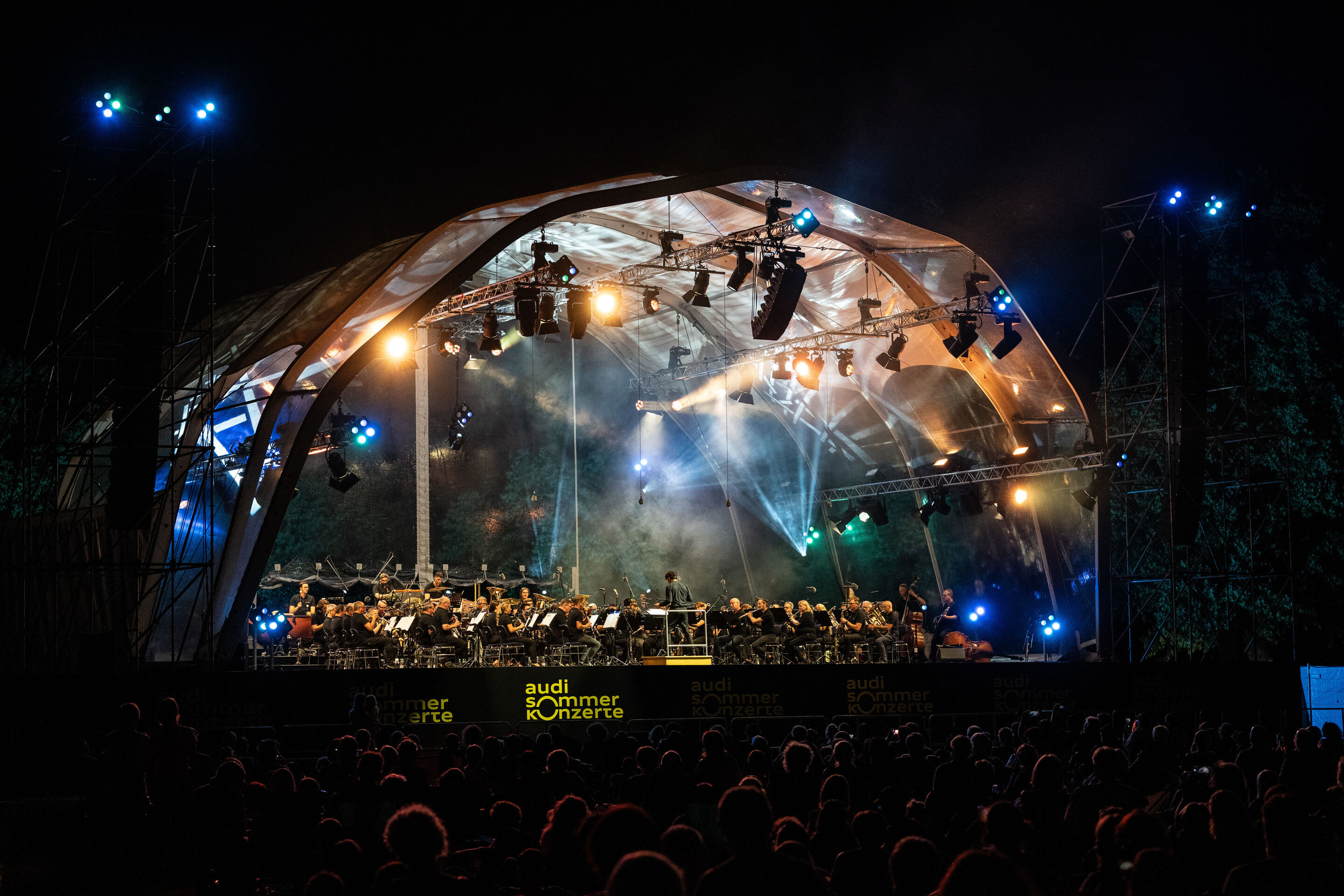 Die Audi Bläserphilharmonie beim Eröffnungskonzert/Klassik Open Air im Klenzepark - Die Audi Bläserphilharmonie unter der Leitung von Mathias Wehr musiziert unter der beleuchteten Bühnenkonstruktion im Klenzepark während des Klassik Open Airs der Audi...
