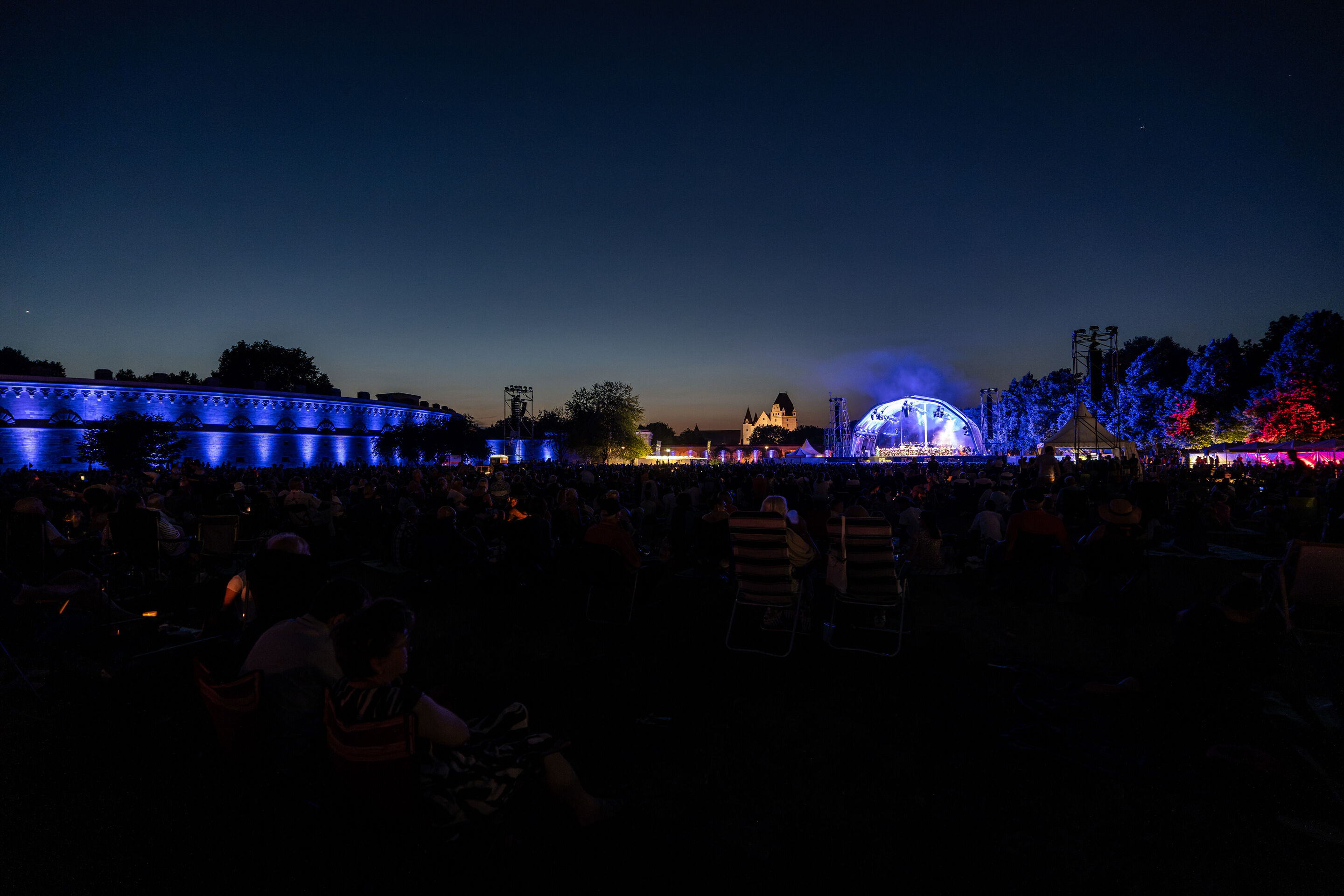 Die Audi Bläserphilharmonie beim Eröffnungskonzert/Klassik Open Air im Klenzepark - Blick über die Wiese  des Klenzeparks bei Nacht auf die beleuchtete Open-Air-Bühne; links ist das blau illuminierte Reduit Tilly zu sehen, im Hintergrund die Silhouette...