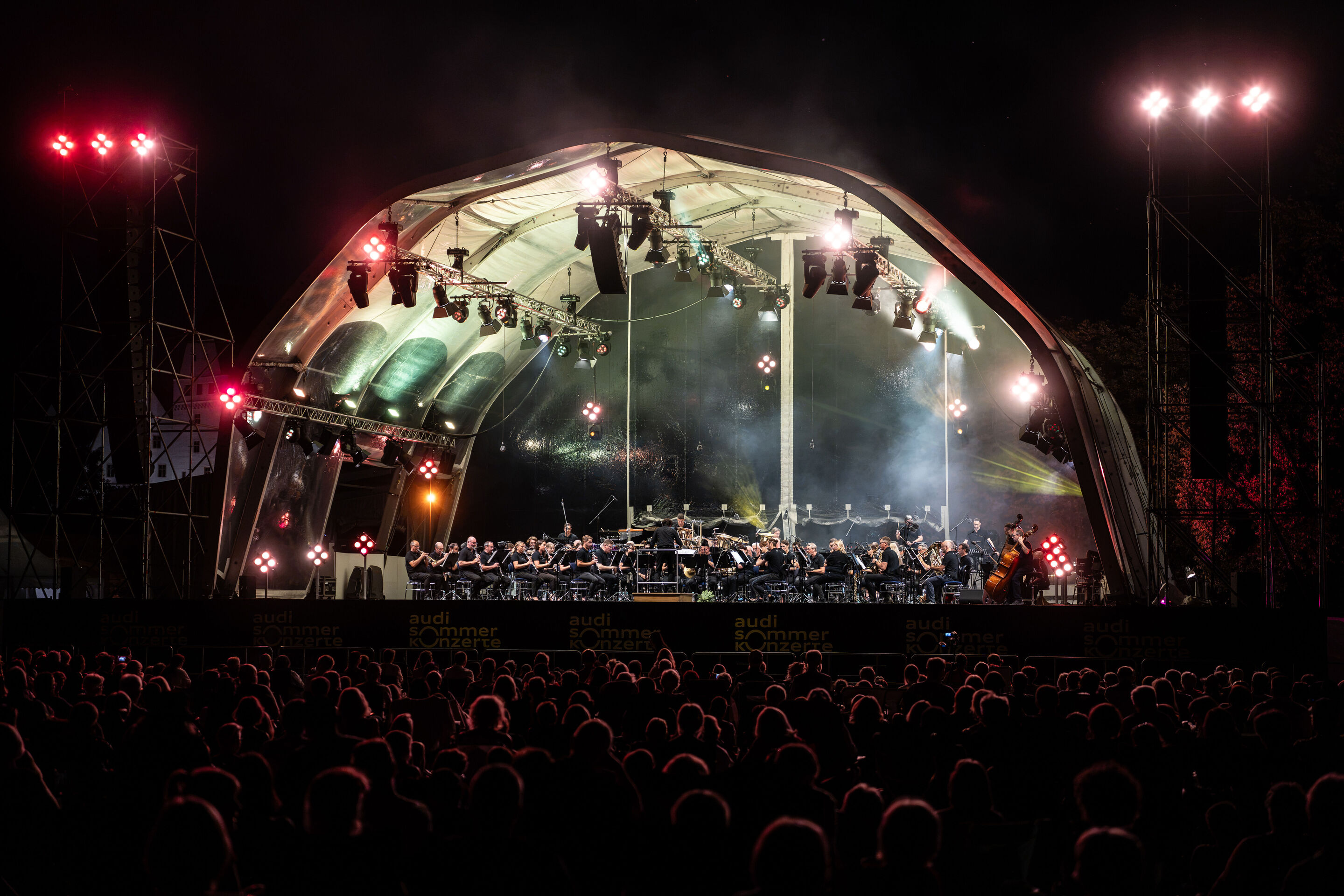 Die Audi Bläserphilharmonie beim Eröffnungskonzert/Klassik Open Air im Klenzepark - Blick aus dem Publikum auf die beleuchtete Open-Air-Bühne im Klenzepark  bei dunklem Nachthimmel: Die Audi Bläserphilharmonie musiziert unter der großflächigen Bühnenkonstruktion,...