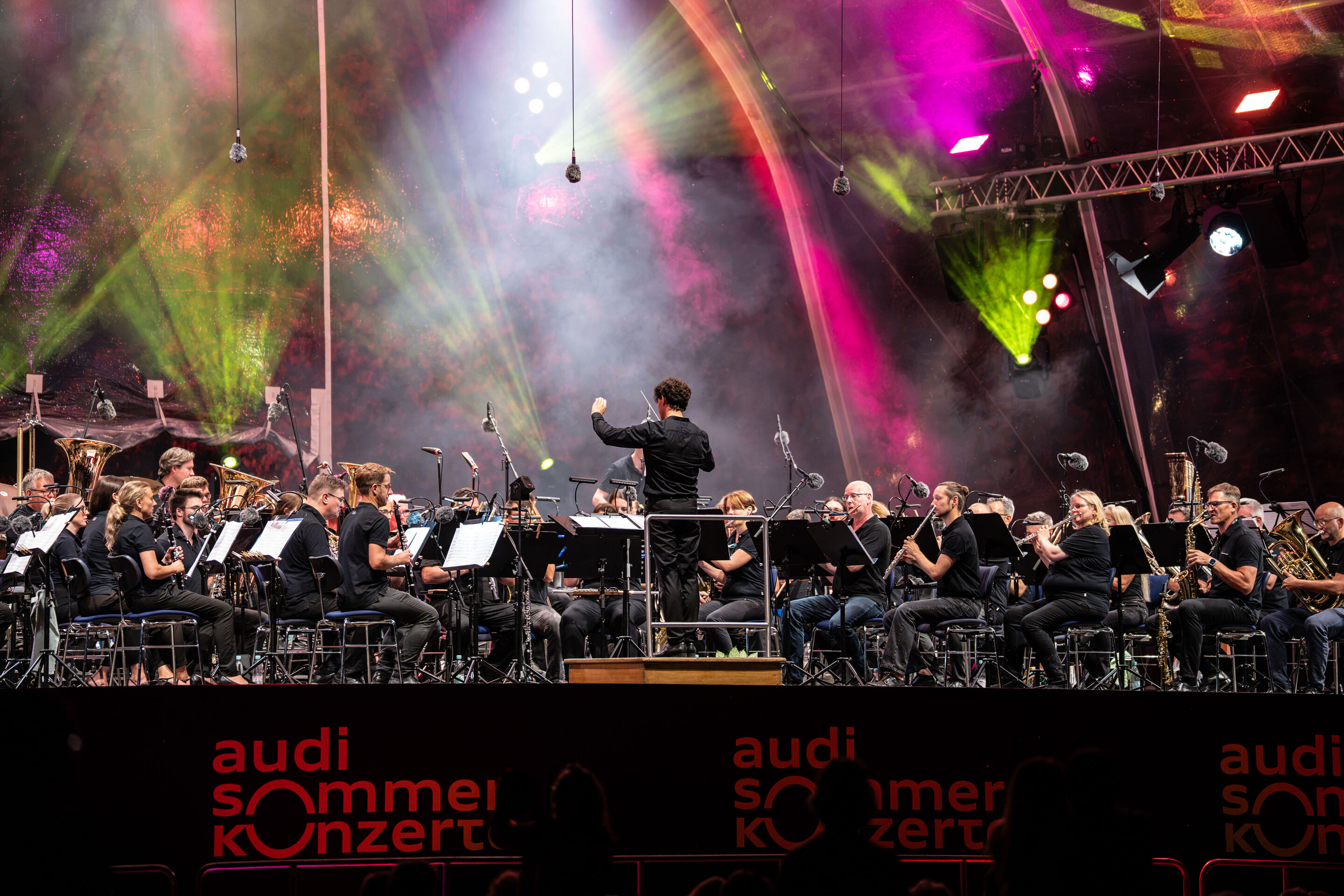 Die Audi Bläserphilharmonie beim Eröffnungskonzert/Klassik Open Air im Klenzepark - Blick aus dem Publikum auf die Bühne des Klassik Open Airs im Klenzepark 2026: Die Audi Bläserphilharmonie musiziert unter der beleuchteten Open-Air-Bühnenkonstruktion,...