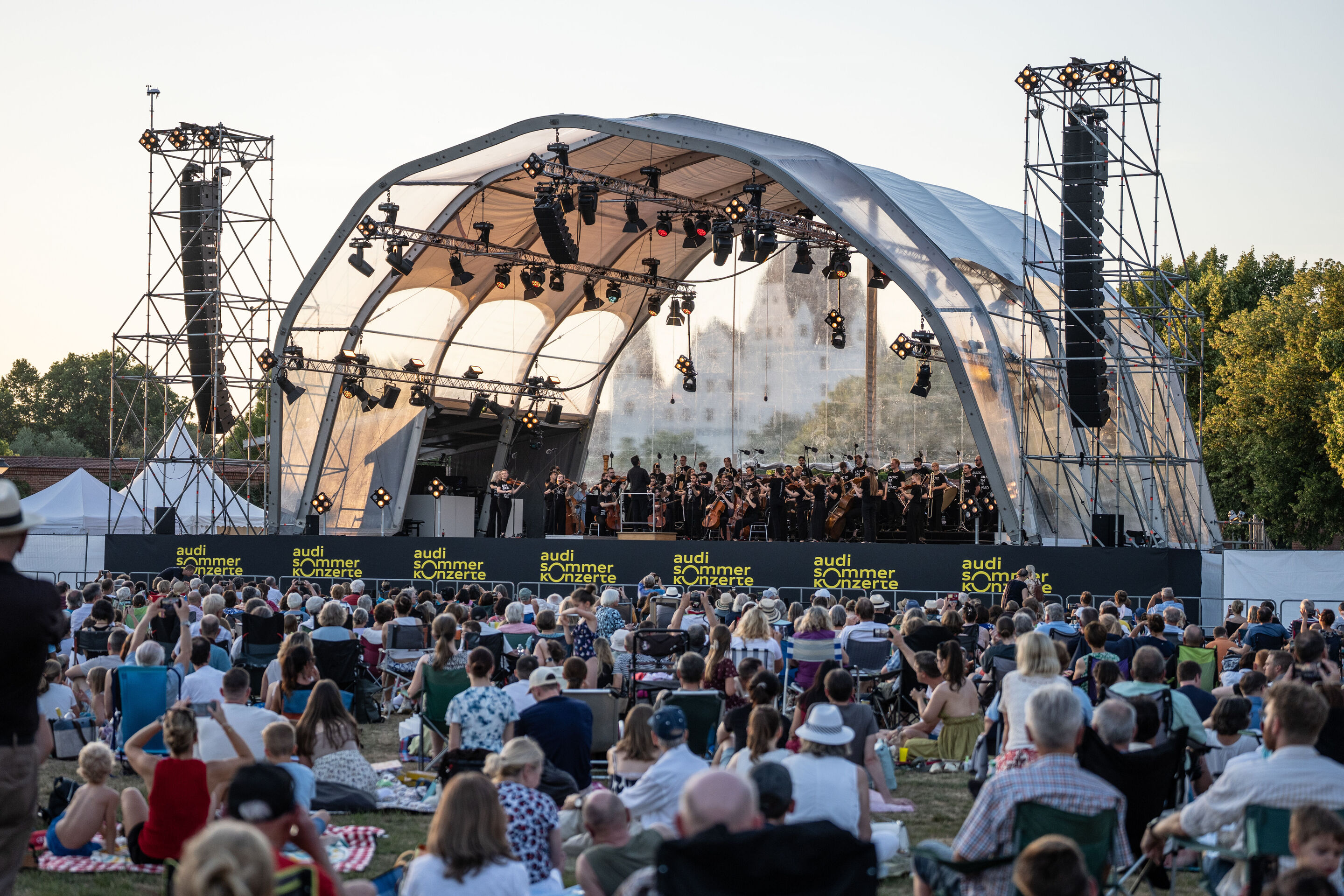 Baltic Sea Philharmonic beim Klassik Open Air im Klenzepark - Blick aus dem Publikum auf die Bühne des Klassik Open Airs im Klenzepark 2026: Das Baltic Sea Philharmonic musiziert unter der Open-Air-Bühnenkonstruktion, während die Besucherinnen und Besucher...