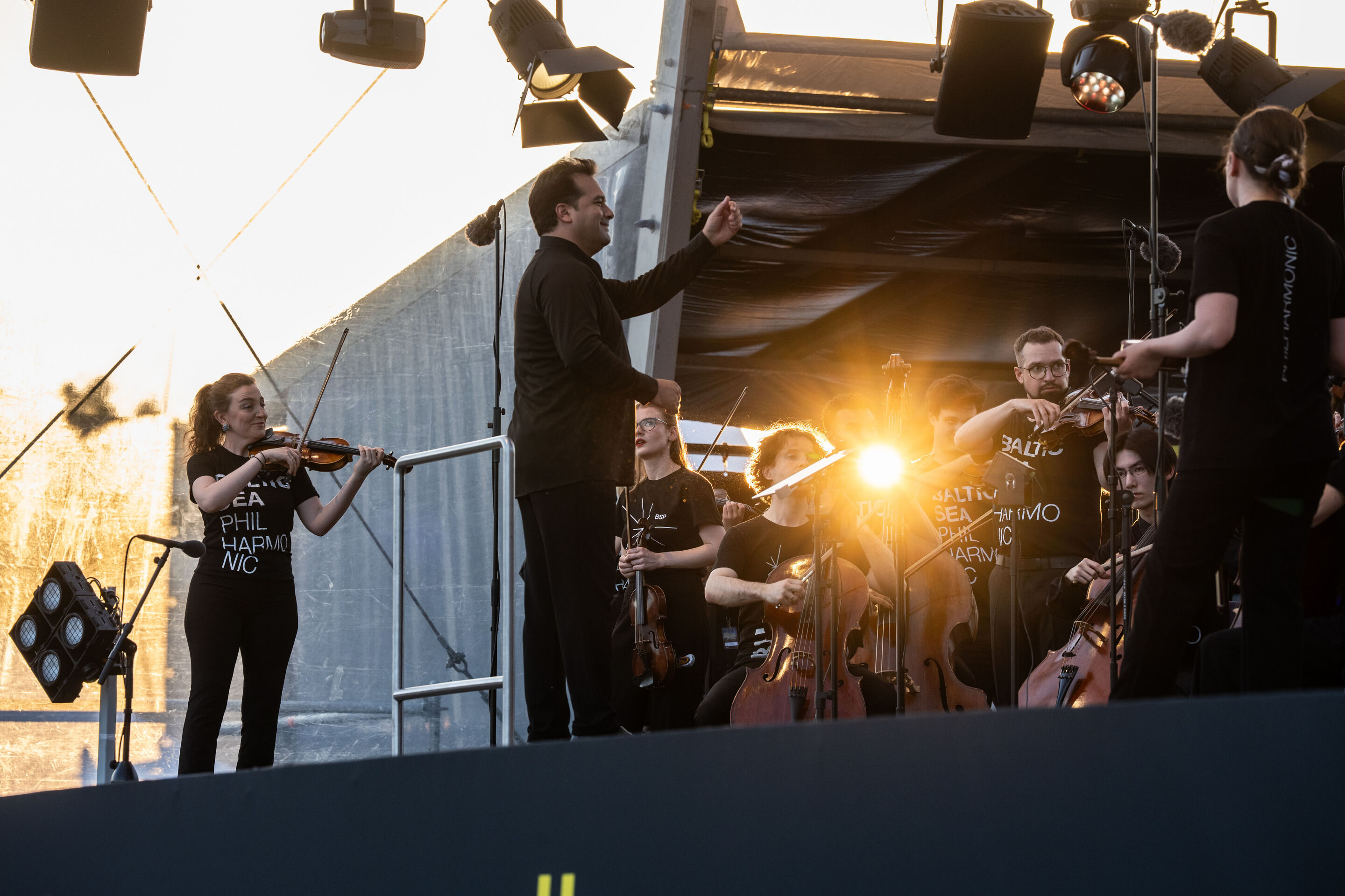 Baltic Sea Philharmonic beim Klassik Open Air im Klenzepark - Dirigent Robert Treviño dirigiert das Baltic Sea Philharmonic beim Klassik Open-Air im Klenzepark 2026, während die Musikerinnen und Musiker mit ihren Instrumenten auf der Bühne vor dem Licht...