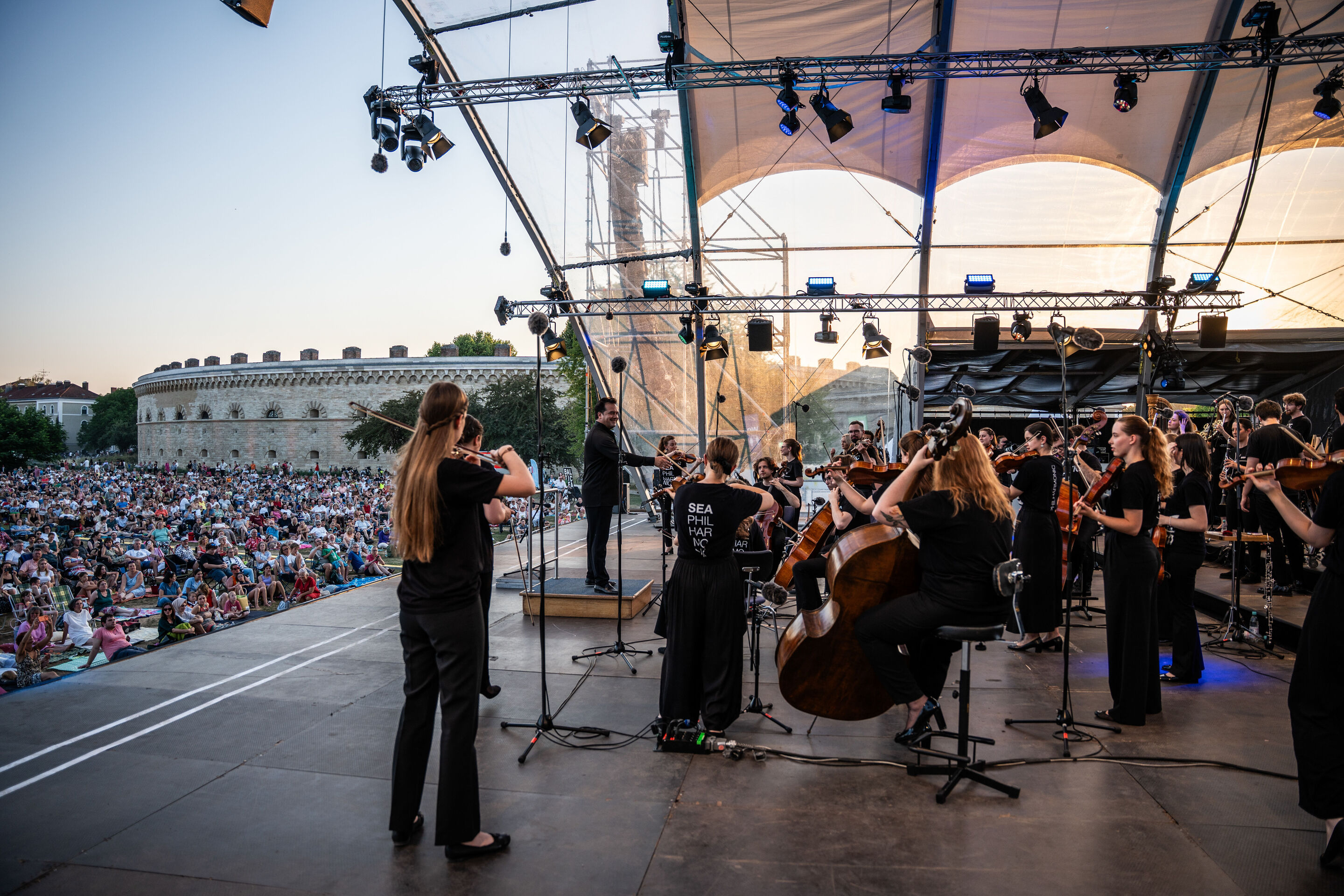 Baltic Sea Philharmonic beim Klassik Open Air im Klenzepark - Blick von der Bühne des Klassik Open-Airs im Klenzepark 2026 auf das Baltic Sea Philharmonic und Dirigent Robert Treviño sowie auf das Publikum vor der Bühne; im Hintergrund ist das Reduit...