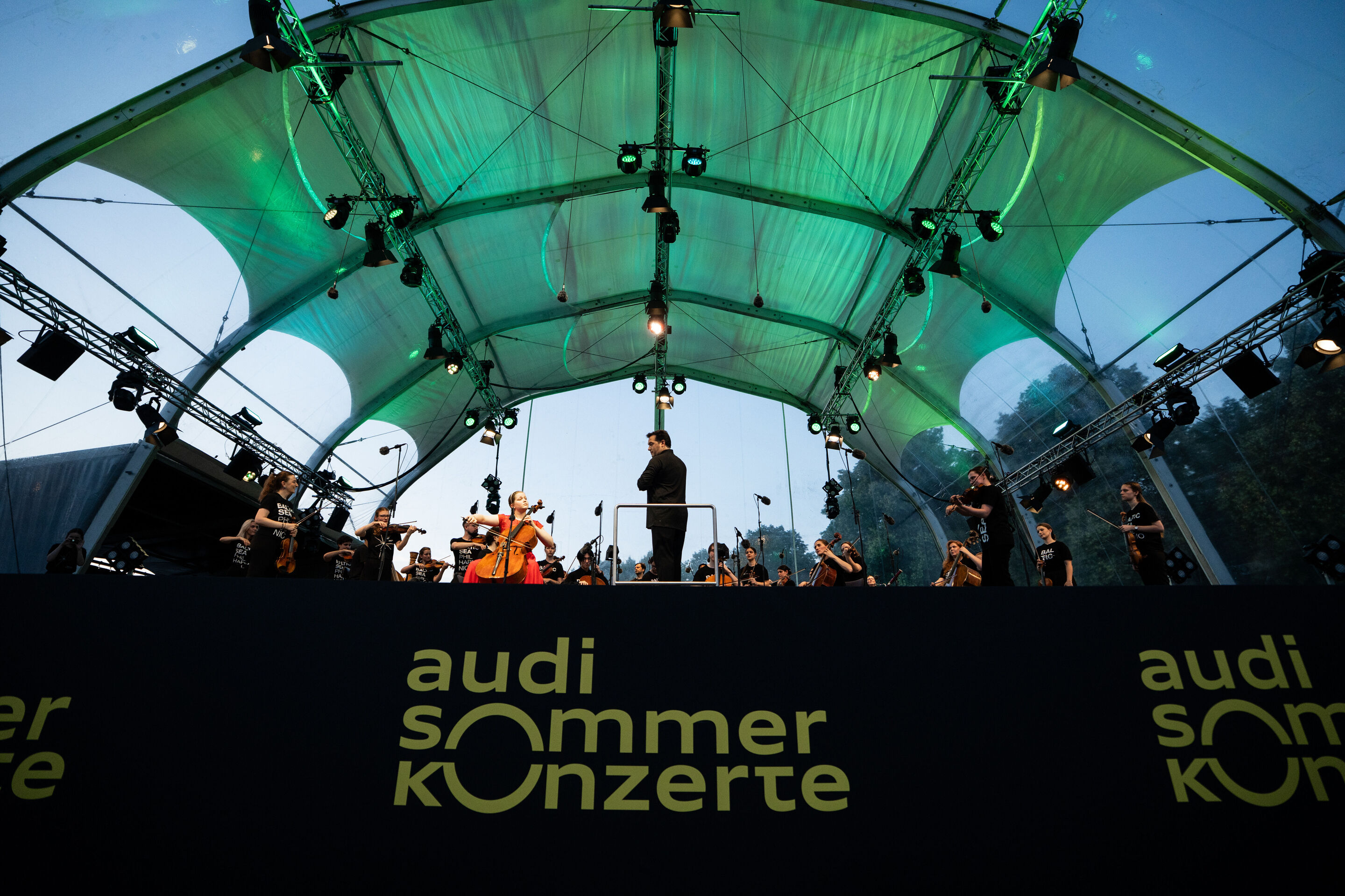 Baltic Sea Philharmonic beim Klassik Open Air im Klenzepark - Blick aus dem Publikum auf die Bühne des Klassik Open-Airs im Klenzepark 2026: Unter der grün beleuchteten Bühnenkonstruktion stehen die Musikerinnen und Musiker des Baltic Sea Philharmonic...