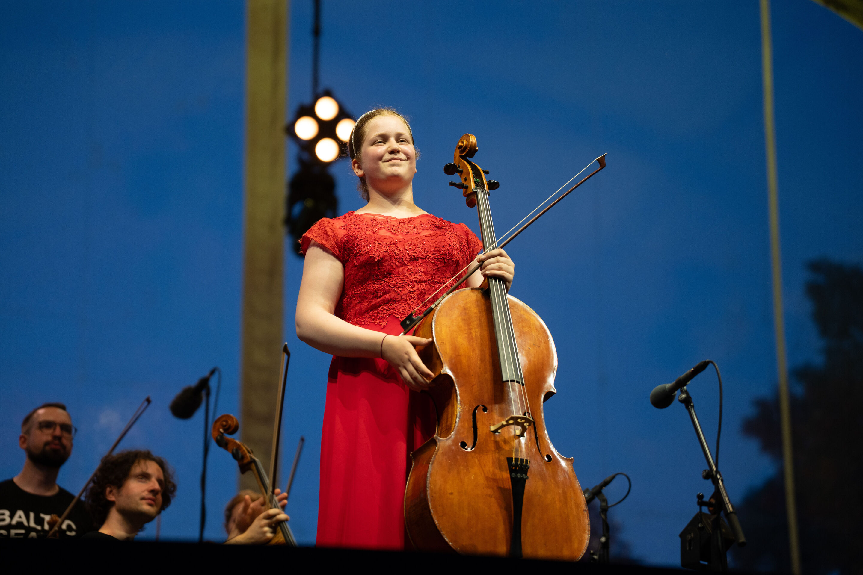 Baltic Sea Philharmonic beim Klassik Open Air im Klenzepark - Die Cellistin Charlotte Melkonian steht mit ihrem Violoncello auf der Bühne des Klassik Open-Airs im Klenzepark 2026, während Mitglieder des Baltic Sea Philharmonic im Hintergrund zu sehen...