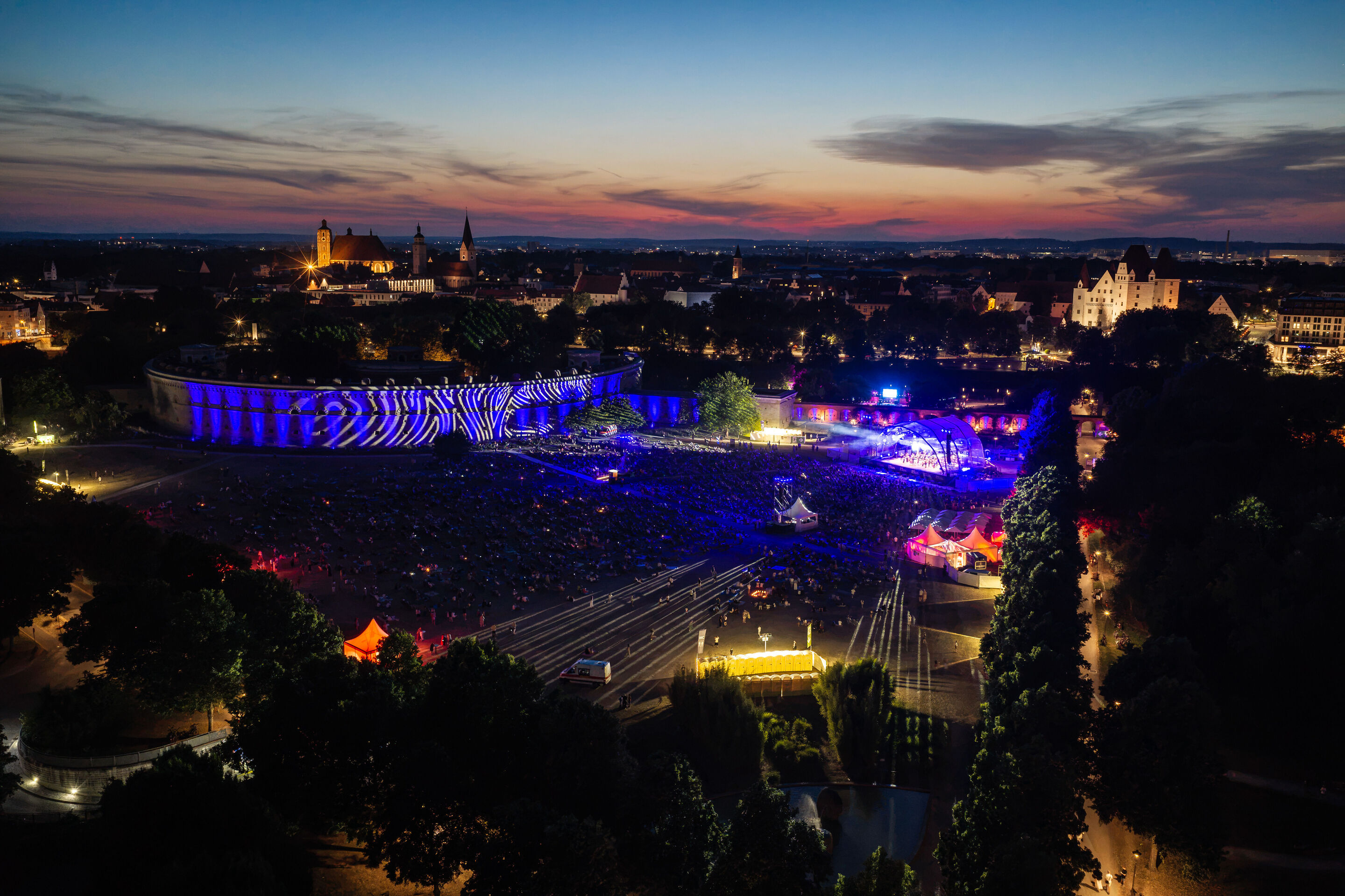 Baltic Sea Philharmonic beim Klassik Open Air im Klenzepark - Vogelperspektive auf das Klassik Open-Air im Klenzepark 2026 mit der blau beleuchteten Konzertbühne, dem illuminierten Reduit Tilly und der Ingolstädter Altstadt im Hintergrund.