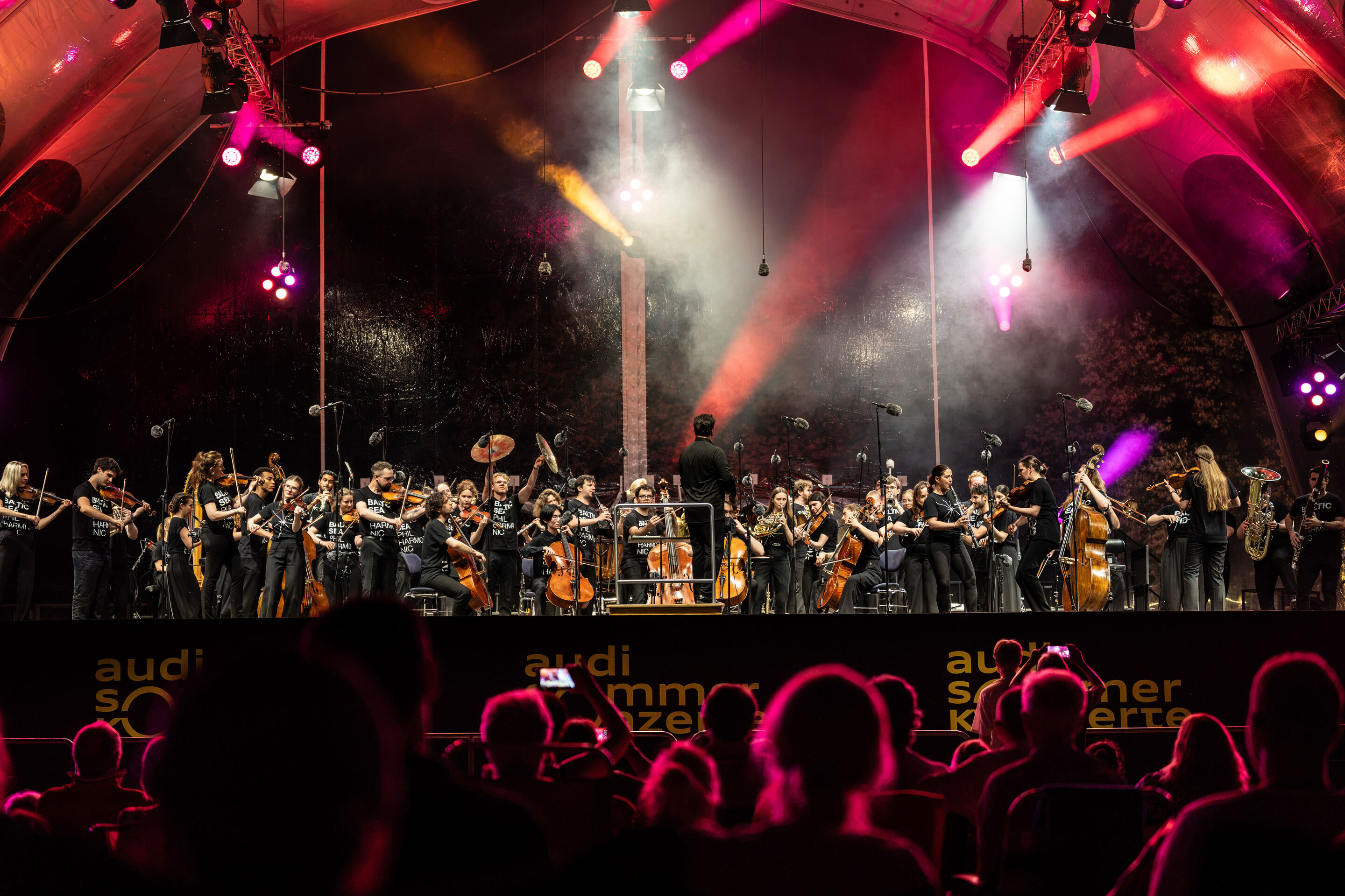 Baltic Sea Philharmonic beim Klassik Open Air im Klenzepark - Blick aus dem Publikum auf die Bühne des Klassik Open-Airs im Klenzepark 2026: Das Baltic Sea Philharmonic musiziert unter der beleuchteten Open-Air-Bühnenkonstruktion, während farbige Lichtinstallationen...