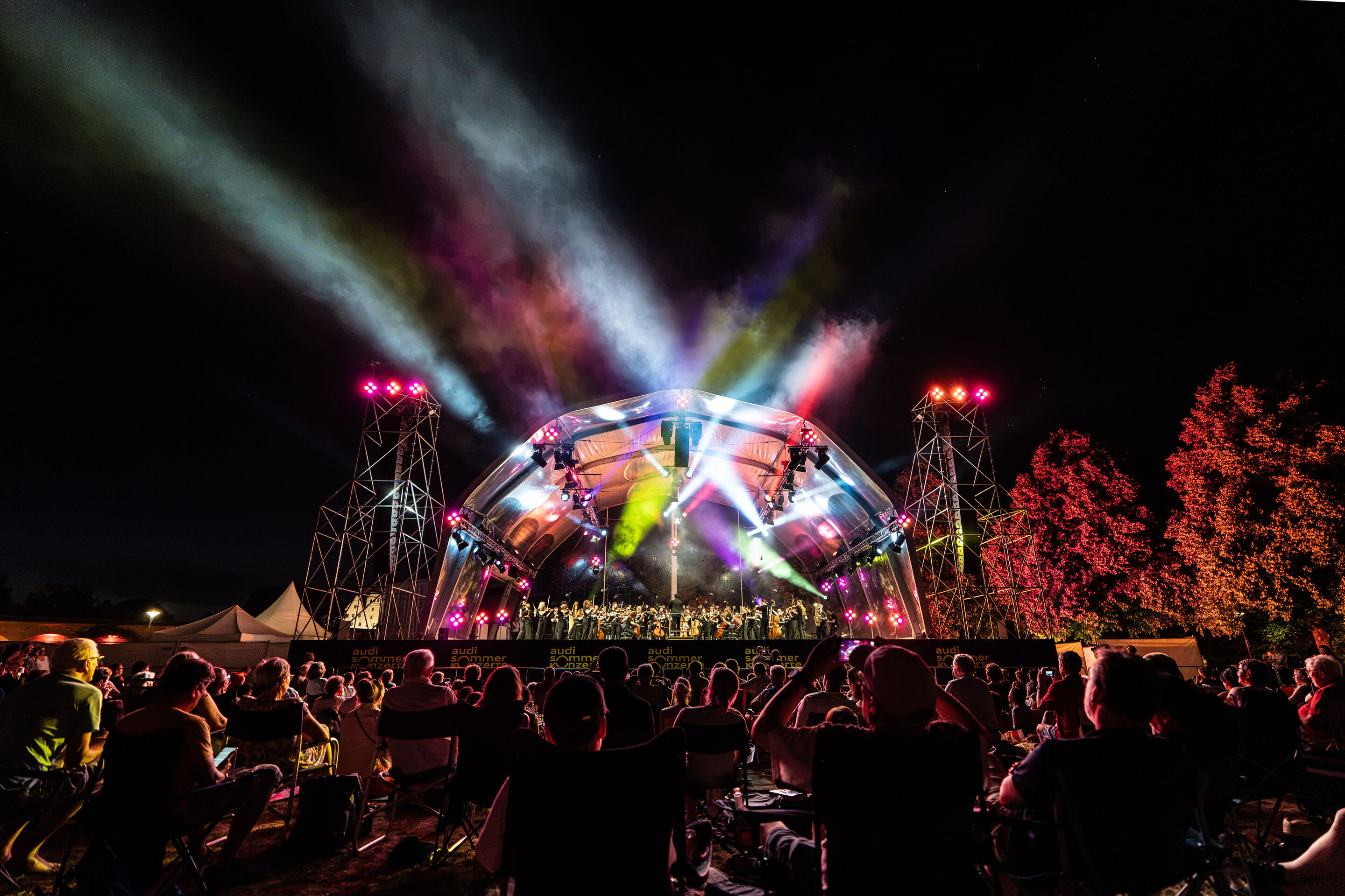 Baltic Sea Philharmonic beim Klassik Open Air im Klenzepark - Blick aus dem Publikum auf die große Konzertbühne des Klassik Open-Airs im Klenzepark 2026, die von farbigen Lichtinstallationen ausgeleuchtet wird, während die Besucherinnen und Besucher das...