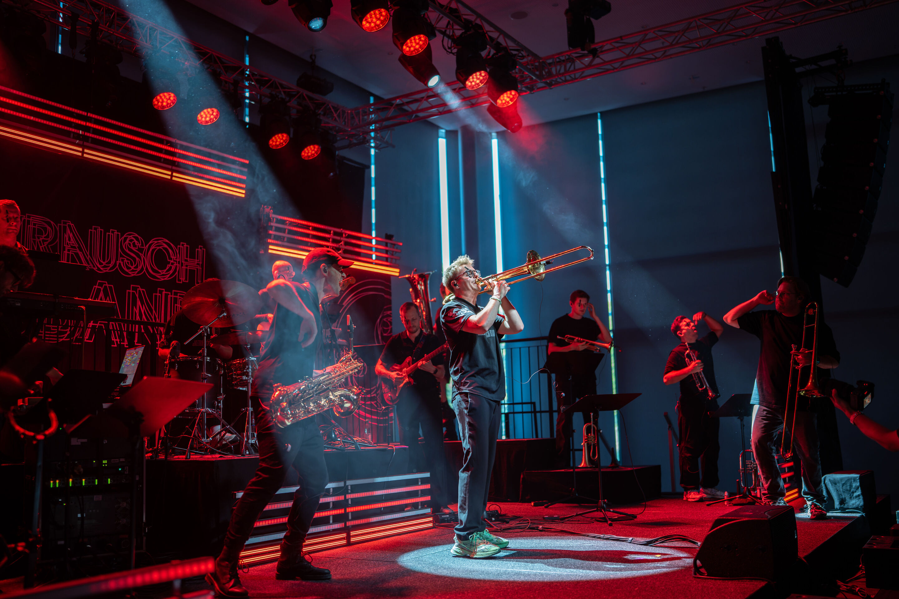 Audi Summer concert: Jazzrausch Bigband performing “Verdi’s Breakdown” - View of the red-lit stage in the Audi H6 building featuring several members of the Jazzrausch Bigband. Brass players are visible in the foreground, while additional musicians perform...