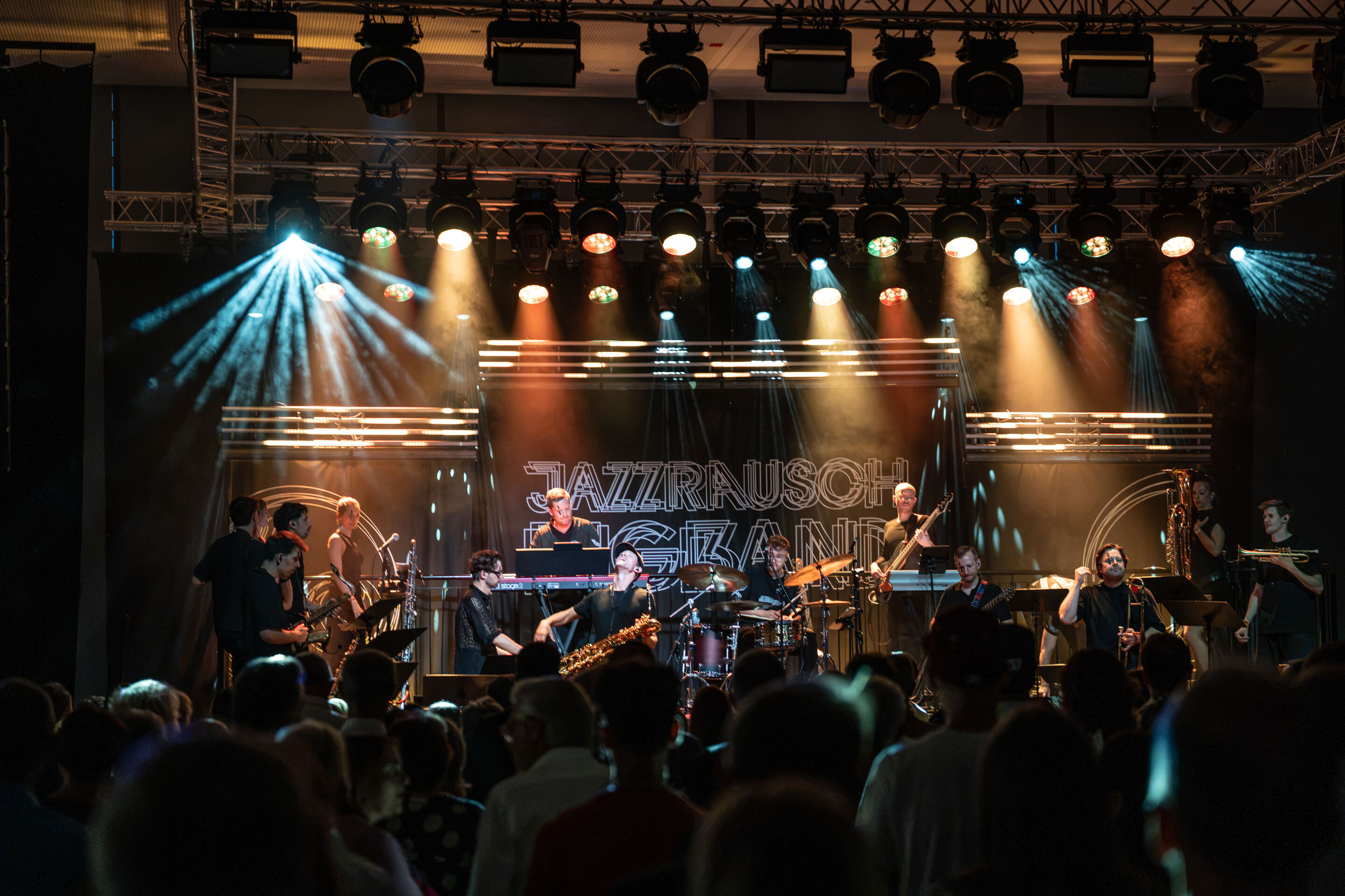 Audi Summer concert: Jazzrausch Bigband performing “Verdi’s Breakdown” - View from the audience toward the stage in the Audi H6 building: The Jazzrausch Bigband performs on the multicolored illuminated stage while audience members watch the concert from...