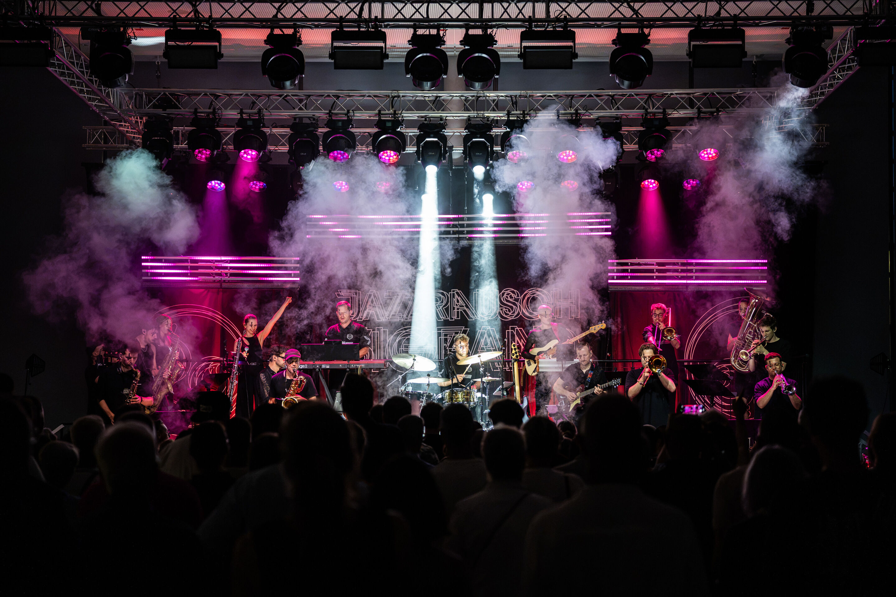 Audi Summer concert: Jazzrausch Bigband performing “Verdi’s Breakdown” - The Jazzrausch Bigband performs on the pink and white illuminated stage in the Audi H6 building. Wisps of stage fog and beams of light fill the stage, while audience members watch...