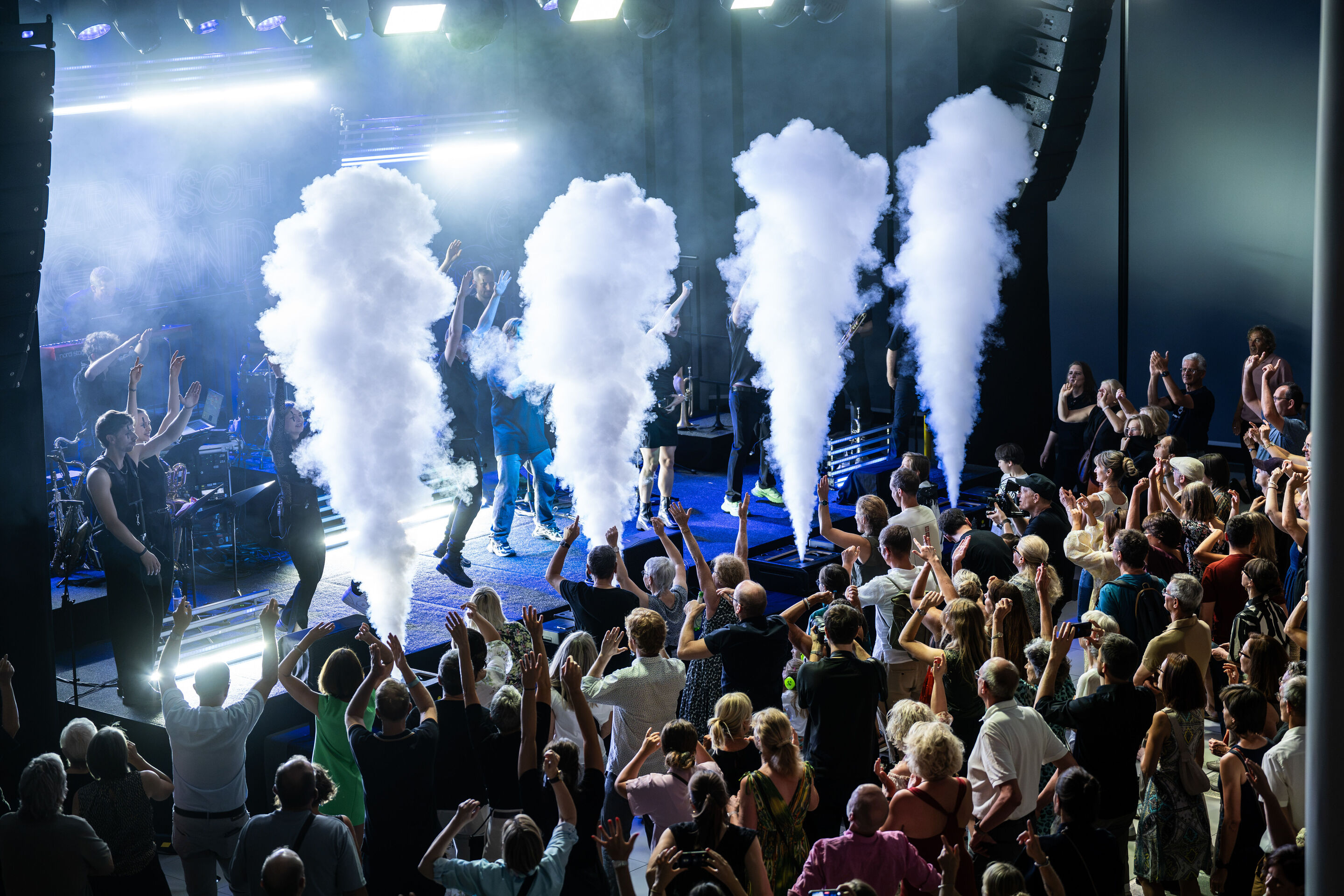 Audi Summer concert: Jazzrausch Bigband performing “Verdi’s Breakdown” - View from an elevated position of the stage in the Audi H6 building: Several members of the Jazzrausch Bigband perform on the blue- and white-lit stage, while four columns of stage...