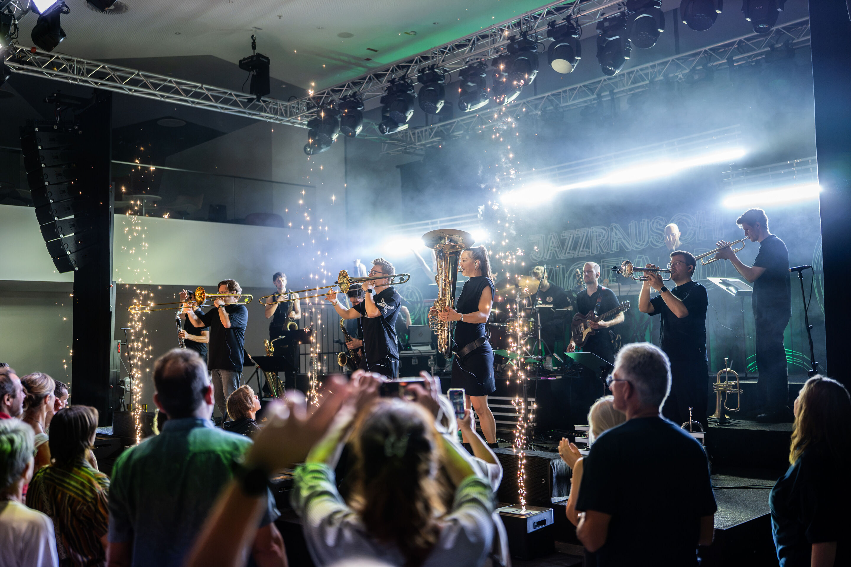 Audi Summer concert: Jazzrausch Bigband performing “Verdi’s Breakdown” - Several members of the Jazzrausch Bigband perform on the white-lit stage in the Audi H6 building. Spark effects are visible in front of the stage, while audience members watch the...