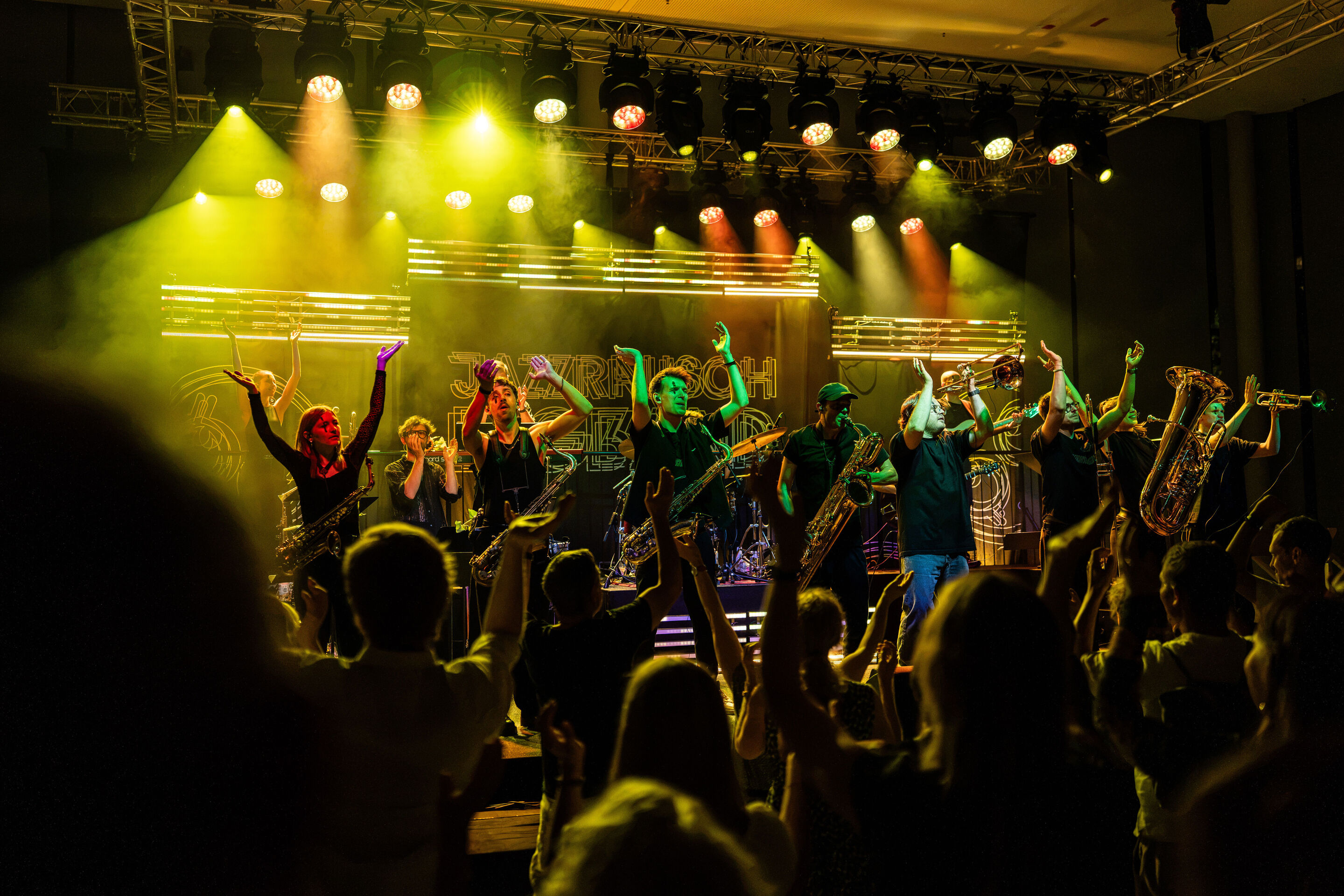 Audi Summer concert: Jazzrausch Bigband performing “Verdi’s Breakdown” - Several members of the Jazzrausch Bigband stand with their instruments on the yellow- and orange-lit stage in the Audi H6 building. They face the audience, while the “Jazzrausch...