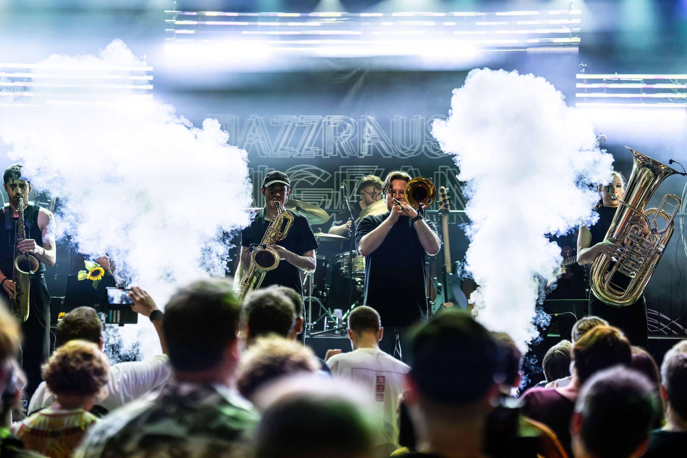 Audi Summer concert: Jazzrausch Bigband performing “Verdi’s Breakdown” - View from the audience toward the stage in the Audi H6 building: Several members of the Jazzrausch Bigband perform under white stage lighting. Thick clouds of stage fog rise to the...