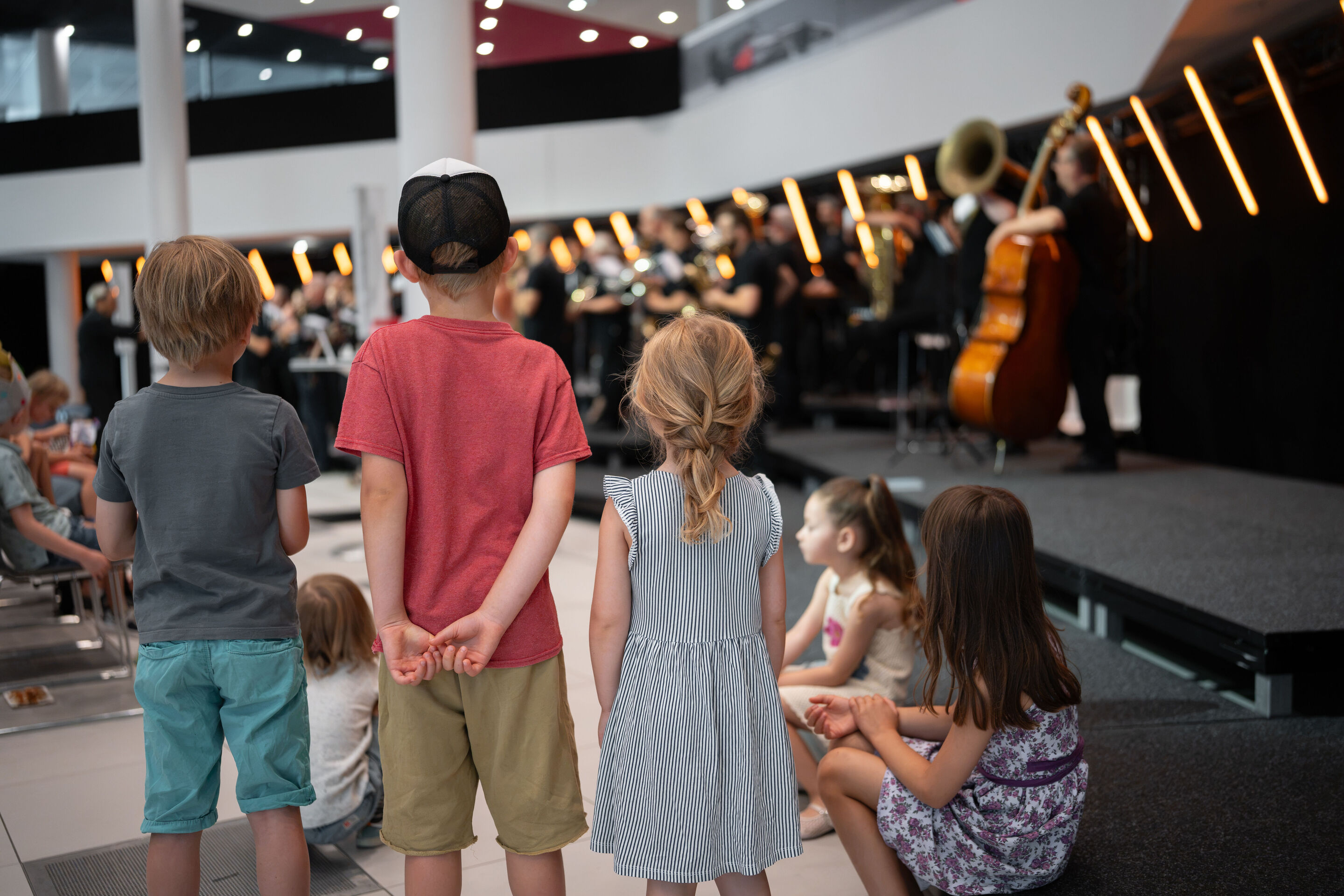 Audi Summer concert: Children’s concert “ADUI und der Geschichtenzauber” - Several children are standing and sitting in front of the stage in the Audi H6 building and looking toward the Audi Bläserphilharmonie. The ensemble’s musicians can be seen in...
