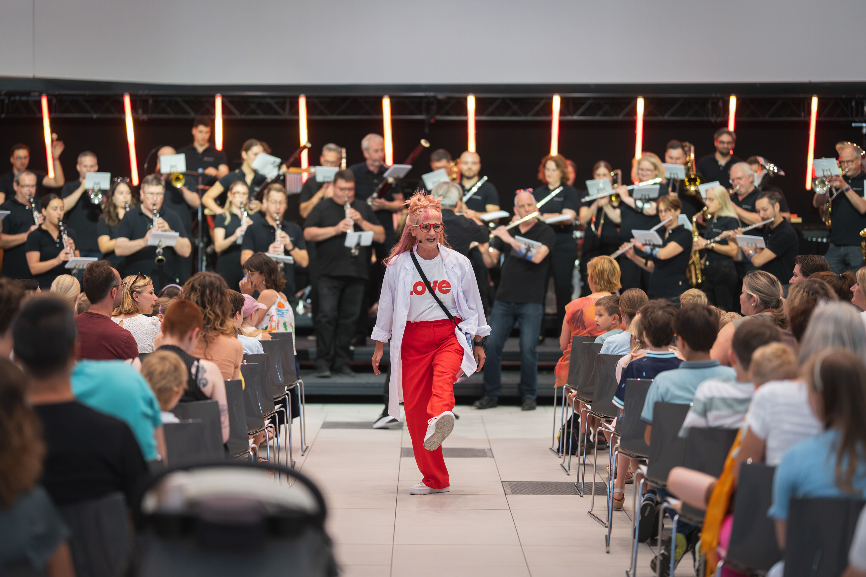 Audi Summer concert: Children’s concert “ADUI und der Geschichtenzauber” - Actress Olivia Wendt moves through the central aisle of the Audi H6 building between the rows of the audience. In the background, the Audi Bläserphilharmonie can be seen on stage.