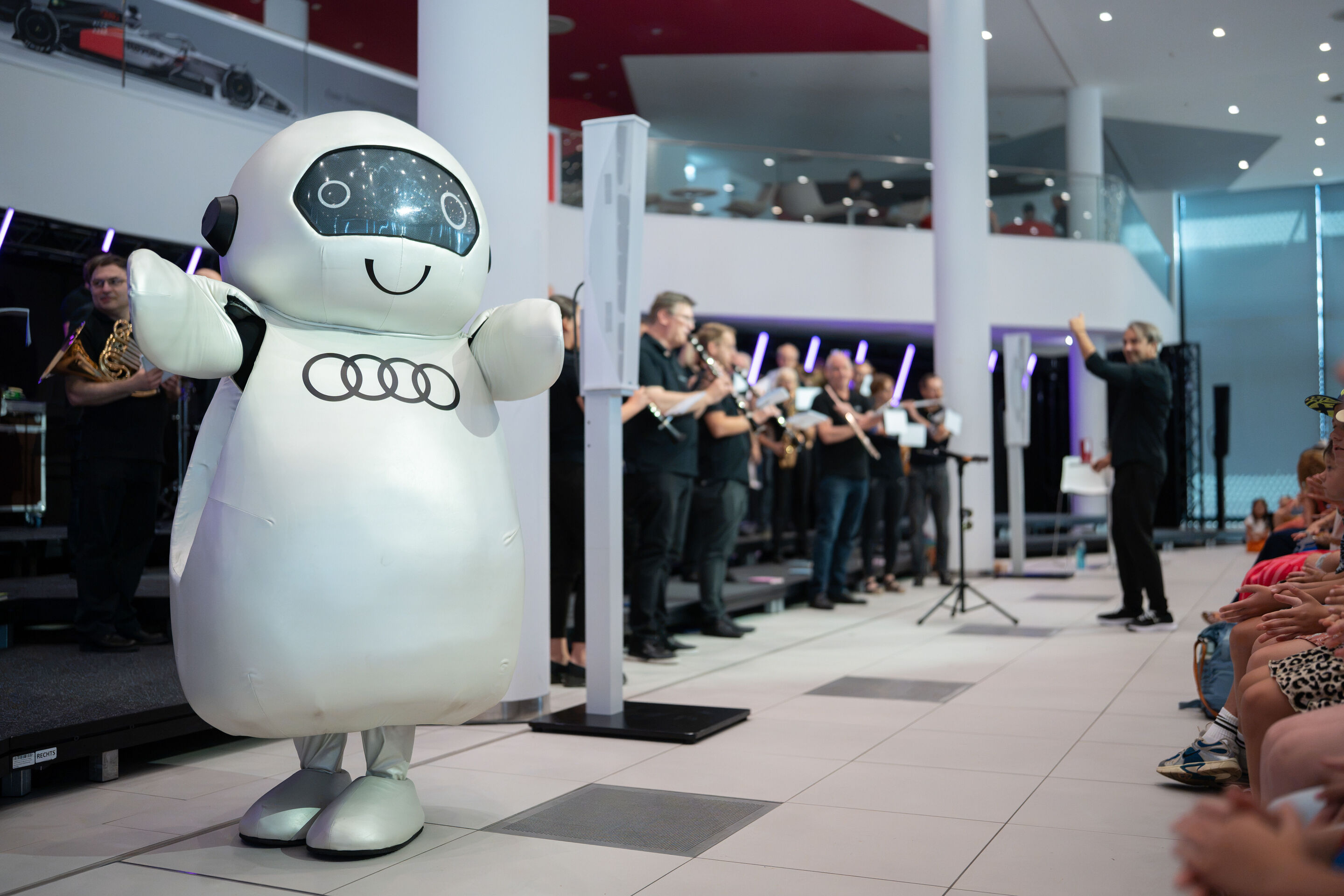 Audi Summer concert: Children’s concert “ADUI und der Geschichtenzauber” - The character ADUI stands in front of the stage in the Audi H6 building with arms outstretched in the foreground of the image. In the background, members of the Audi Bläserphilharmonie...