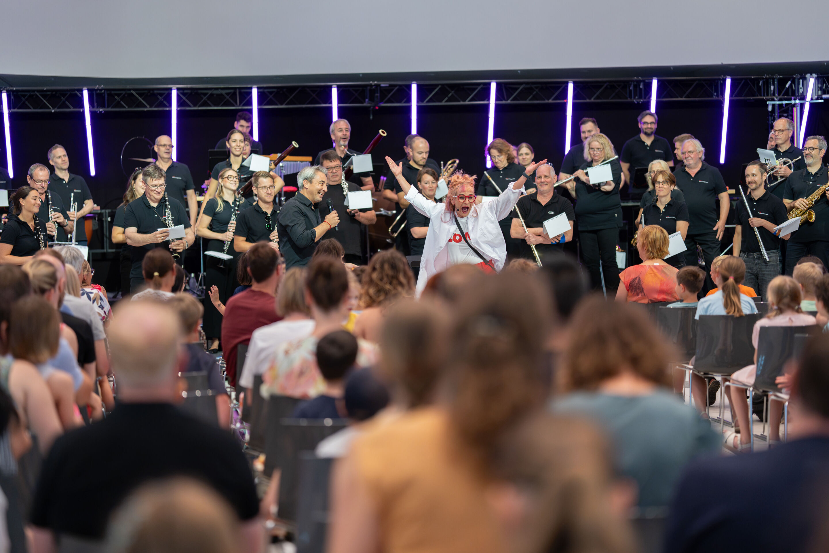 Audi Summer concert: Children’s concert “ADUI und der Geschichtenzauber” - View from the audience toward the performance area in the Audi H6 building: Actress Olivia Wendt stands with her arms outstretched among the members of the Audi Bläserphilharmonie....