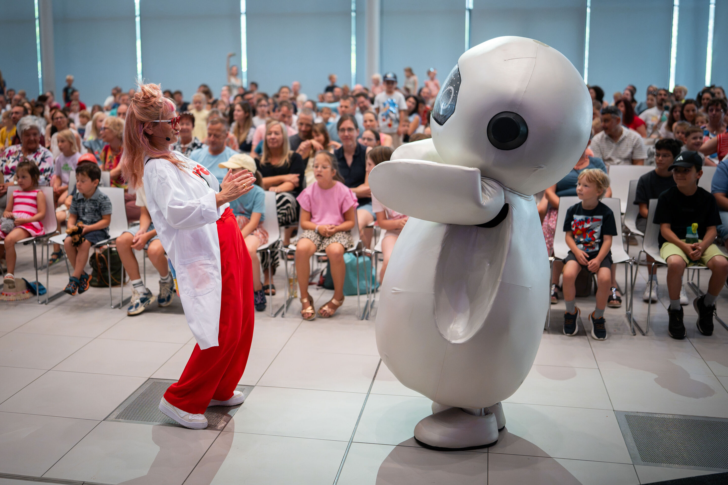 Audi Summer concert: Children’s concert “ADUI und der Geschichtenzauber” - View from the stage toward the audience in the Audi H6 building: Actress Olivia Wendt stands together with the character ADUI in front of the visitors. Olivia Wendt and ADUI face...