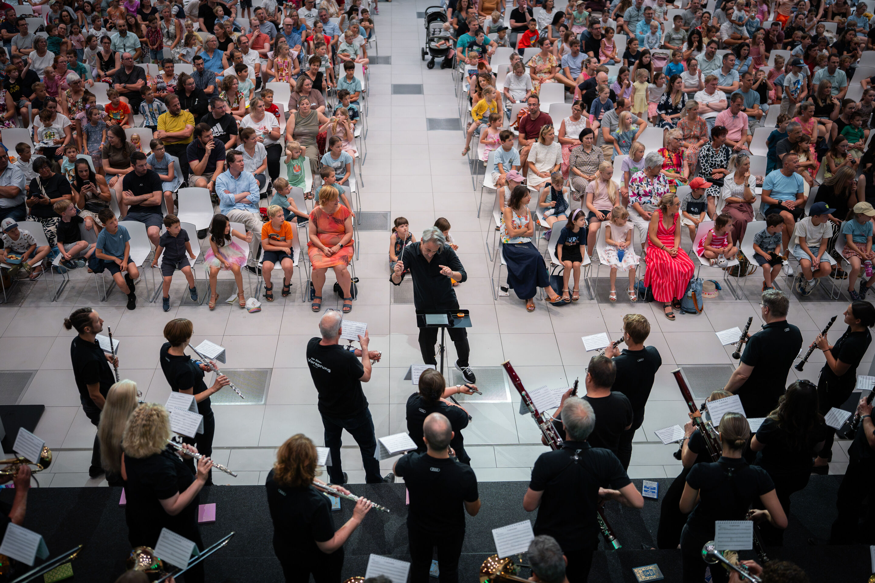 Audi Summer concert: Children’s concert “ADUI und der Geschichtenzauber” - View from an elevated position toward the audience in the Audi H6 building: The Audi Bläserphilharmonie has gathered around conductor and composer Siegmund Andraschek, who is standing...