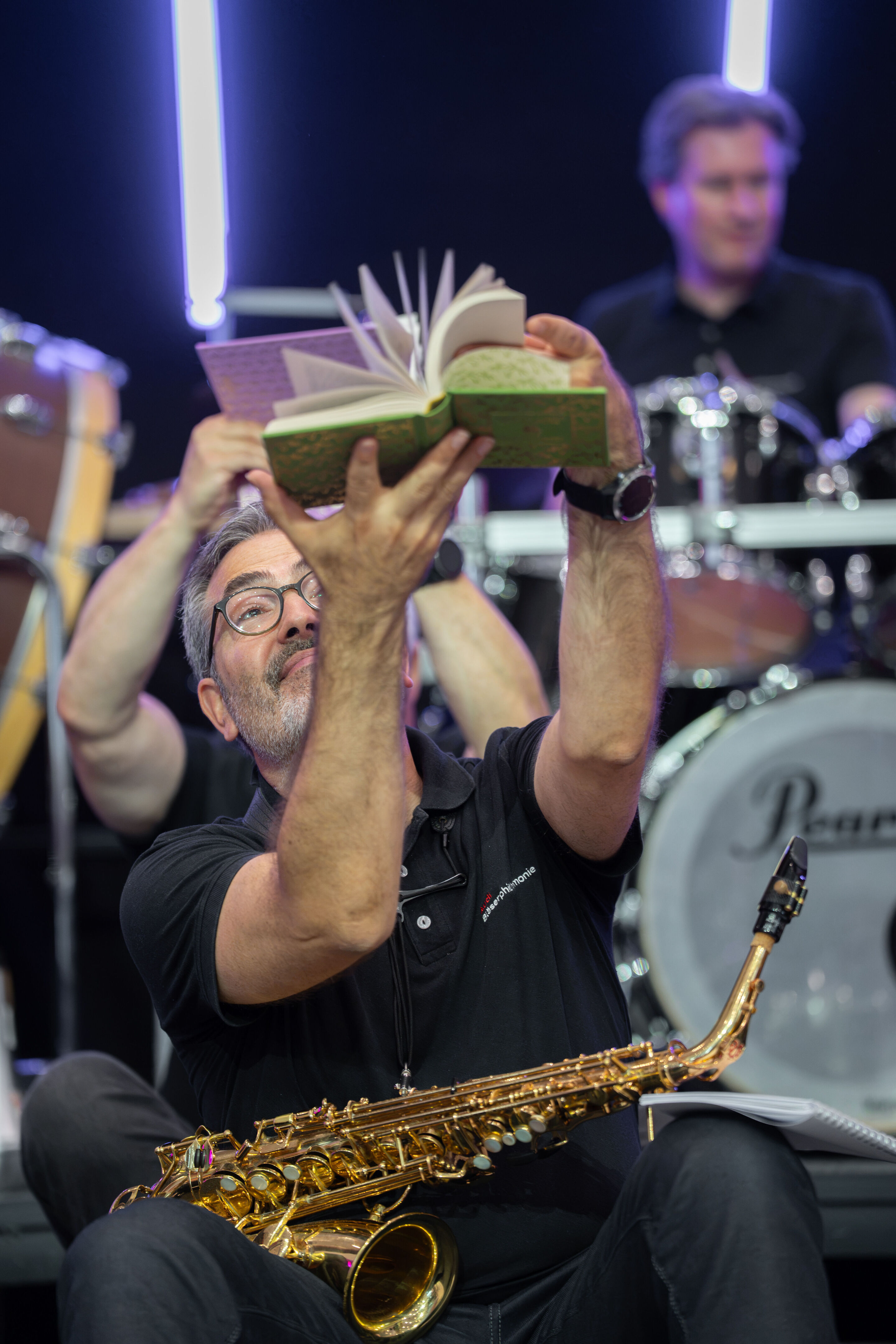 Audi Summer concert: Children’s concert “ADUI und der Geschichtenzauber” - A member of the Audi Bläserphilharmonie sits with a saxophone on the stage in the Audi H6 building and holds an open book above himself. Additional members of the ensemble can...