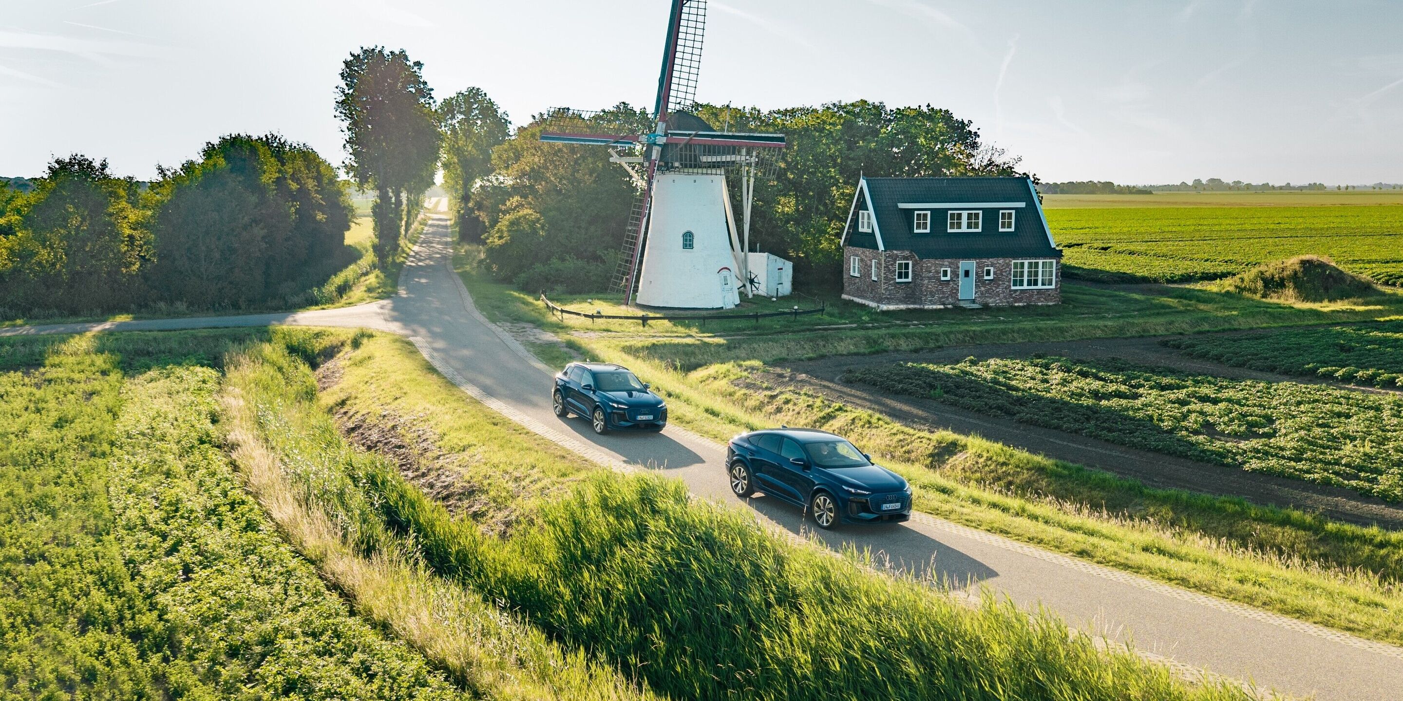 Audi Q6 e-tron Lifecycle Experience - Zwei Audi Q6 e-tron (Ascariblau) fahren hintereinander auf einer schmalen Landstraße durch eine grüne Kulturlandschaft mit Windmühle und Wohnhaus im Hintergrund.