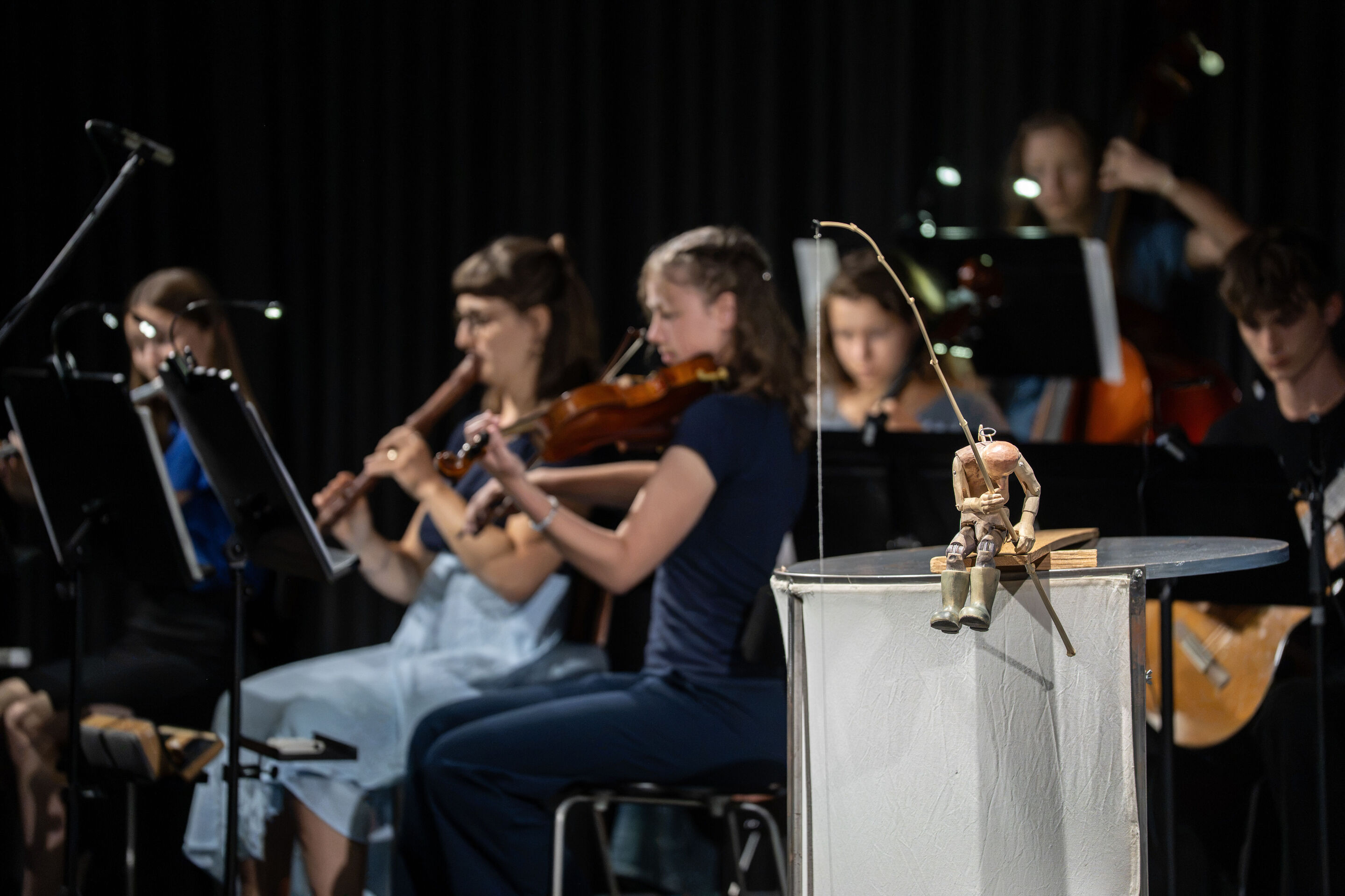 Audi Sommerkonzerte 2026 – Puppentheater „Der Fischer und seine Frau“ - Im Vordergrund der Aufnahme sitzt die Holzfigur des Fischers mit einer Angel auf einem Podest. Dahinter sind Musikerinnen und Musiker des Ensembles der Städtischen Simon-Mayr-Sing-...