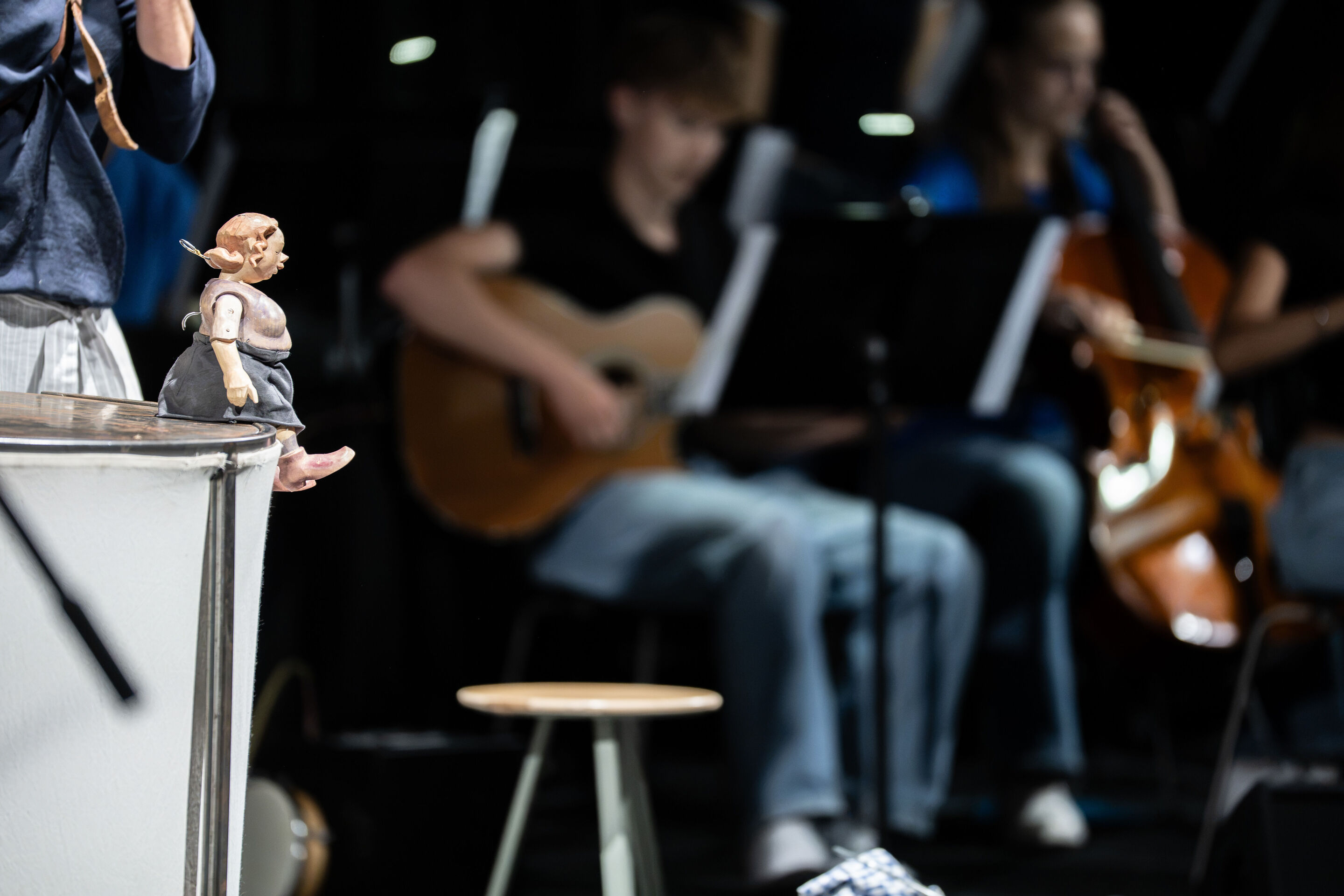Audi Sommerkonzerte 2026 – Puppentheater „Der Fischer und seine Frau“ - Die Holzfigur der Frau des Fischers sitzt auf dem Rand eines Podests im Vordergrund der Aufnahme. Im Hintergrund begleiten Musikerinnen und Musiker des Ensembles der Städtischen Simon-Mayr-Sing-...
