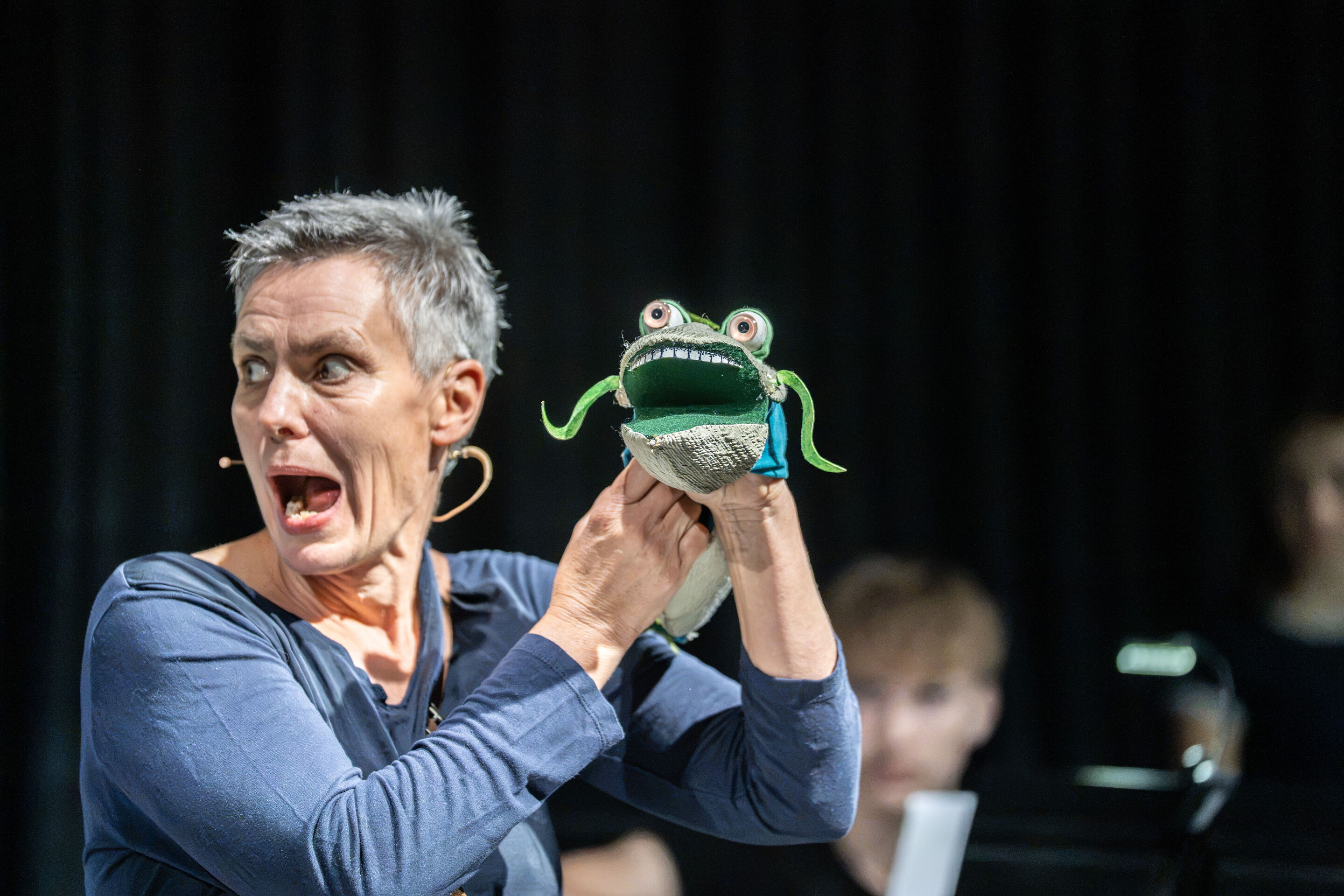 Audi Sommerkonzerte 2026 – Puppentheater „Der Fischer und seine Frau“ - Puppenspielerin Annika Pilstl hält eine Fischfigur auf Schulterhöhe neben ihrem Gesicht. Die Aufnahme zeigt sie mit weit geöffnetem Mund und zur Seite gerichtetem Blick.