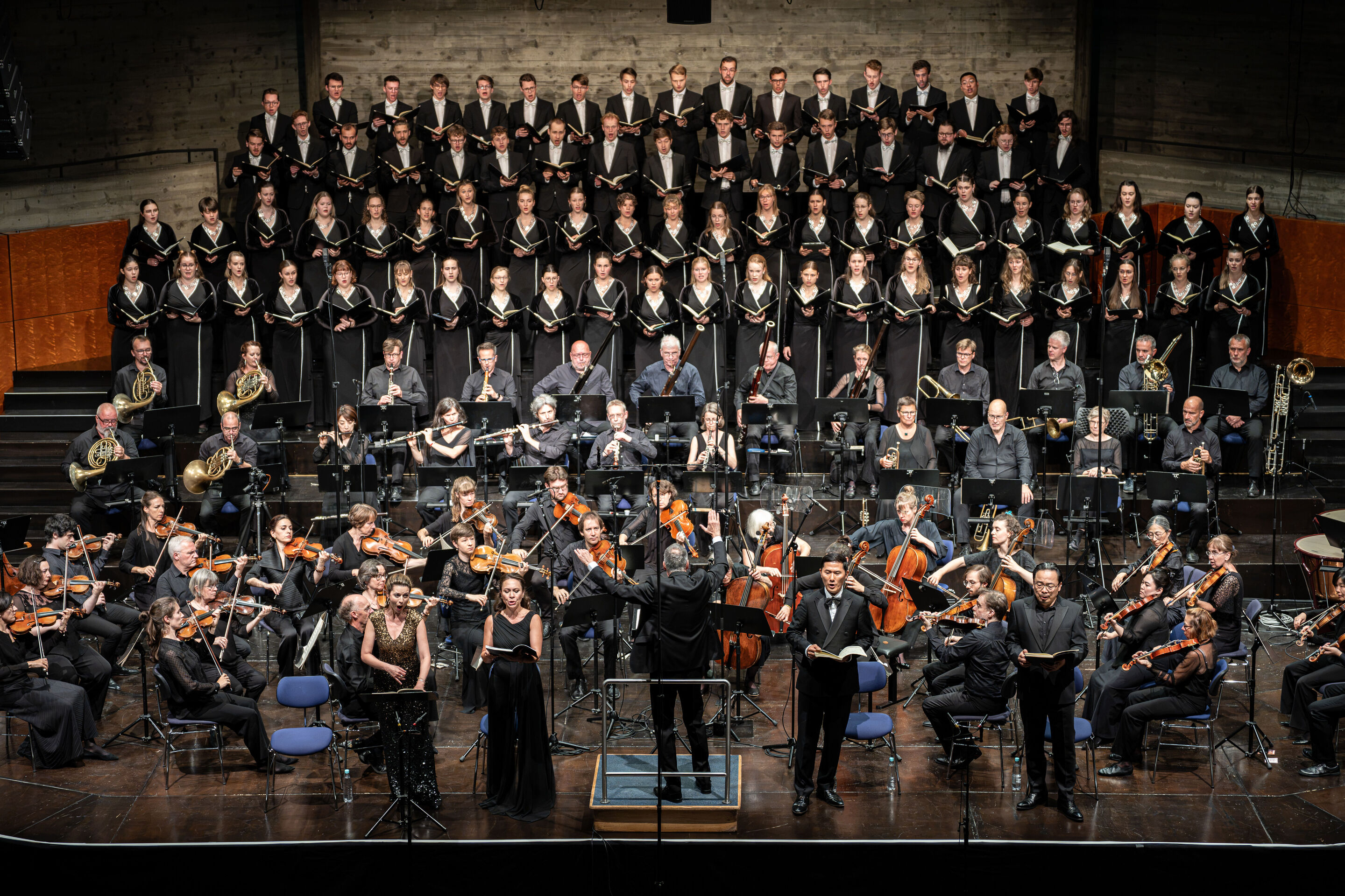 Audi Summer Concerts 2026: Closing concert - View of the stage in the Festsaal Ingolstadt. The Audi Youth Choir Academy stands on tiered choir risers behind the Akademie für Alte Musik Berlin. In the foreground, Martin Steidler conducts the performance...
