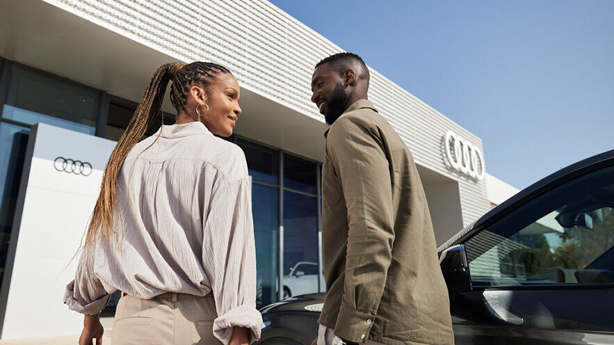 two people - Two people are standing in front of an Audi dealership and a dark-colored car, smiling in the sunny weather.