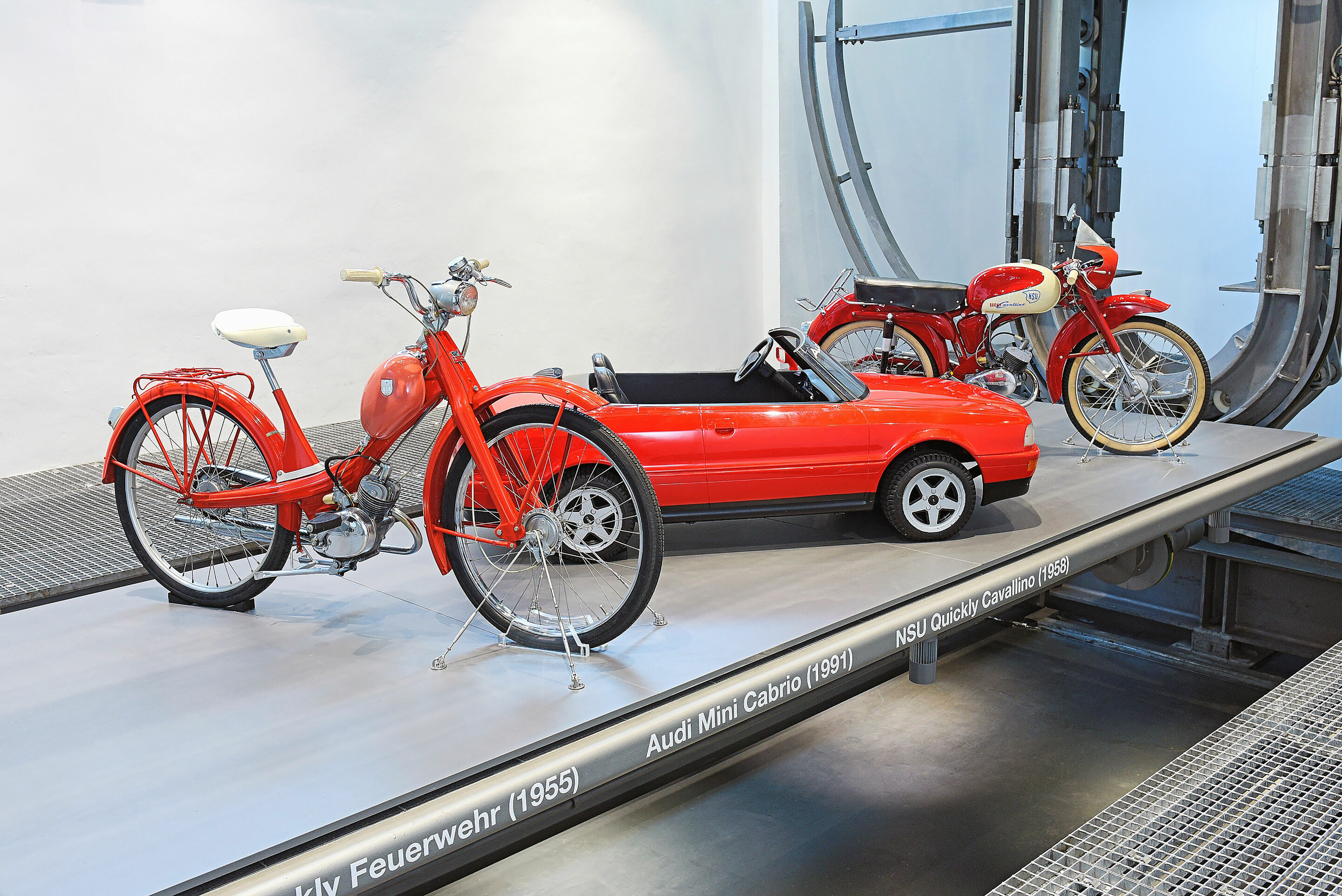 „Variationen in Rot“ im Audi museum mobile - Die Zweiräder NSU Quickly „Feuerwehr“ und „Cavallino“ sowie das Audi Cabriolet im Maßstab 1:2 sind auf einer Plattform präsentiert.